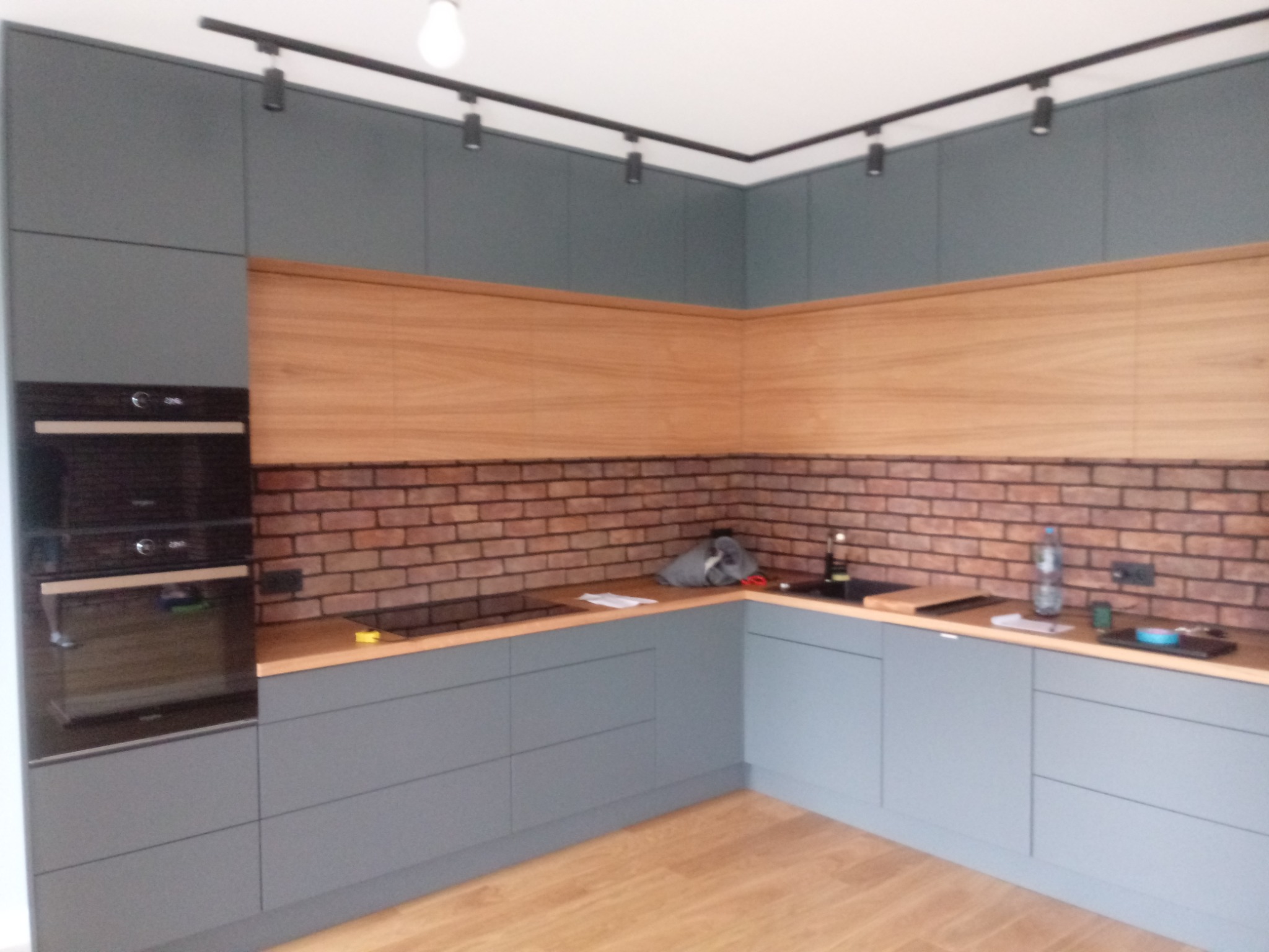 Szara zabudowa kuchenna z drewnianym blatem i ceglaną ścianą, oświetlenie punktowe na suficie.