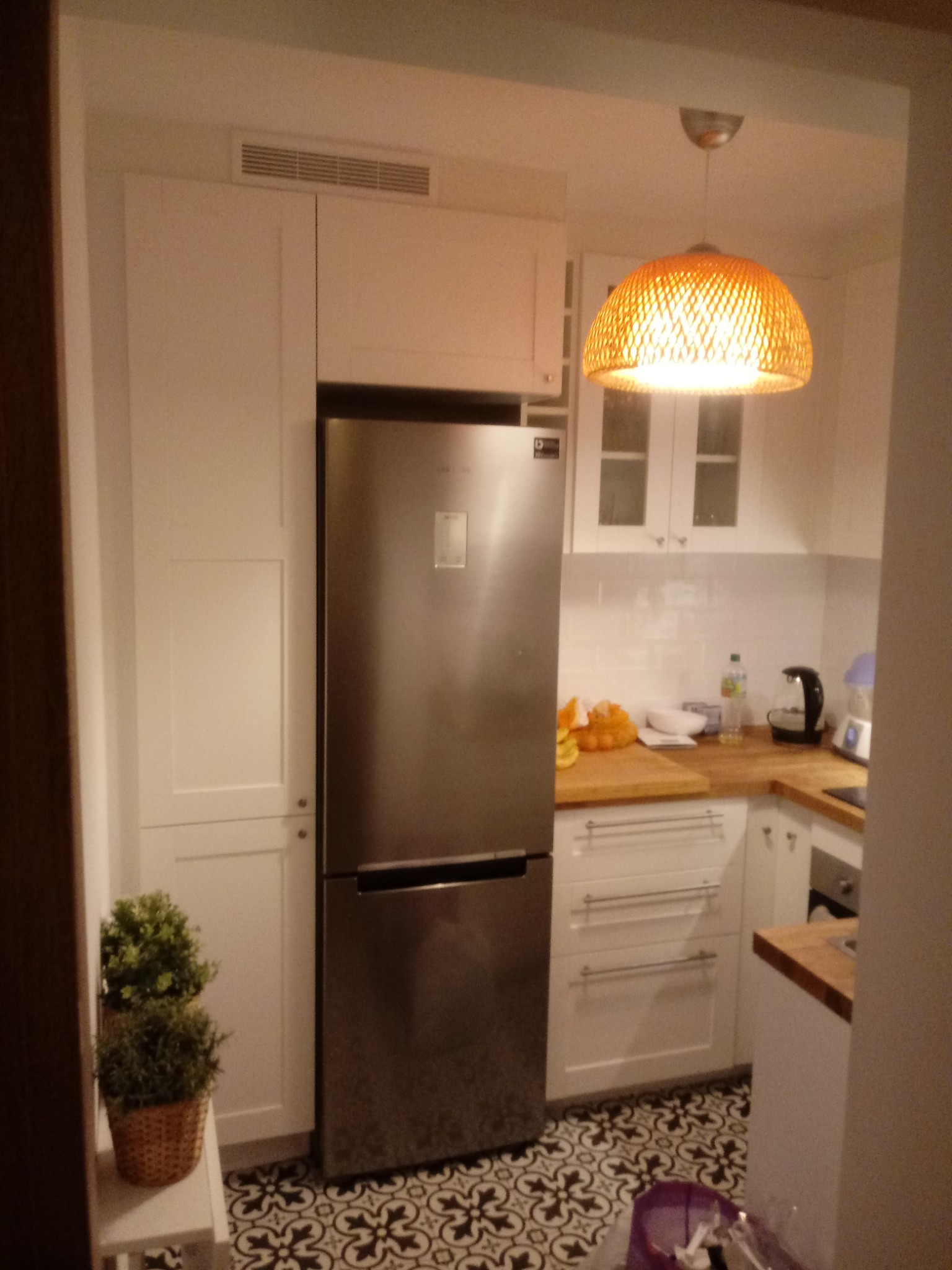 Wnętrze kuchni z białymi szafkami, drewnianym blatem, stalową lodówką i wzorzystą podłogą; widoczna lampa z abażurem z wikliny.