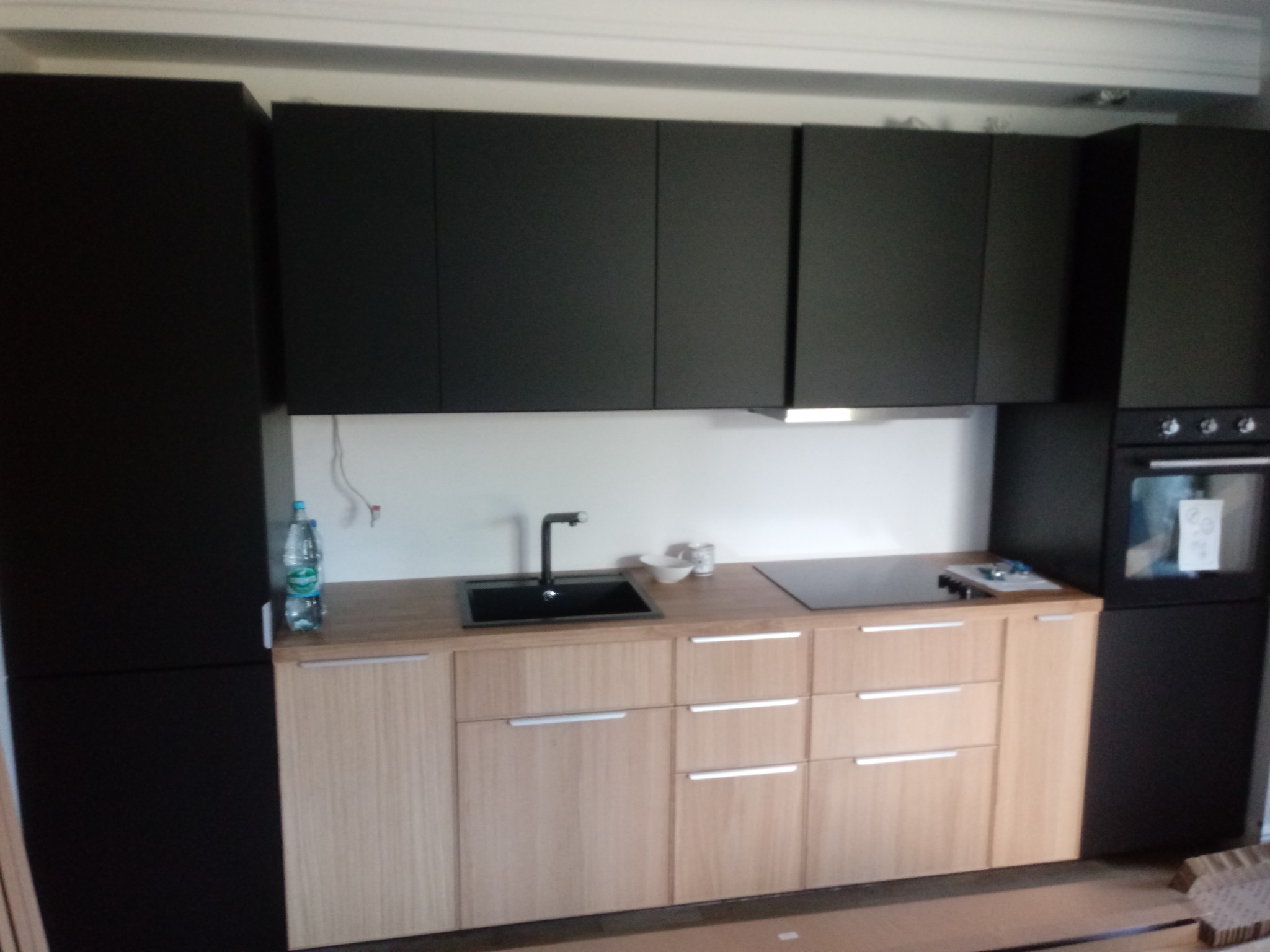 Nowoczesna kuchnia z czarnymi szafkami wiszącymi i jasnymi, drewnianymi szafkami dolnymi, czarny zlew i płyta indukcyjna na drewnianym blacie, butelka wody mineralnej na blacie, elementy montażowe...