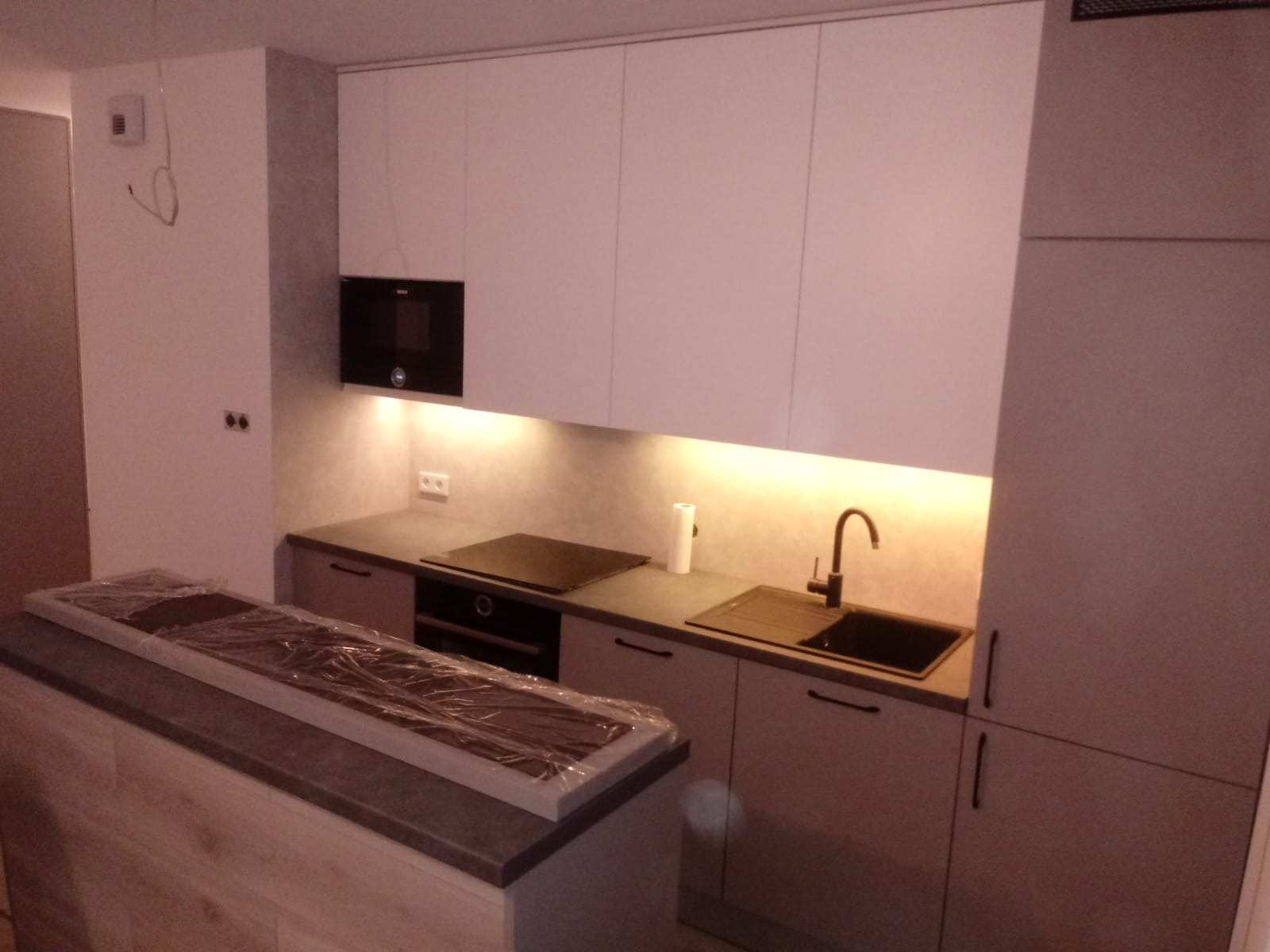 Nowoczesna kuchnia z białymi szafkami, czarną armaturą i blatem wyspowym zabezpieczonym folią ochronną, oświetlenie LED pod szafkami górnymi.