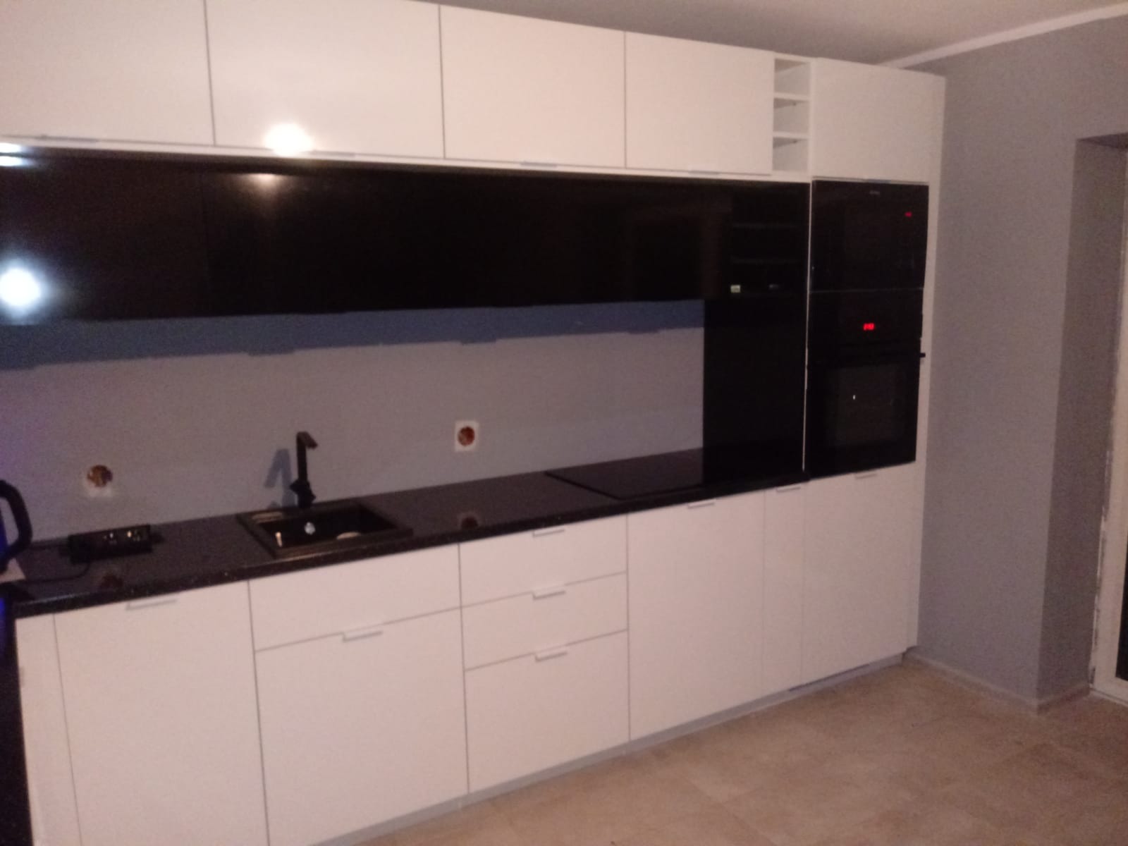 Nowoczesna, biała zabudowa kuchenna z czarnym blatem i panelem ściennym, widoczne otwory na gniazdka przed montażem, zabudowane piekarniki.