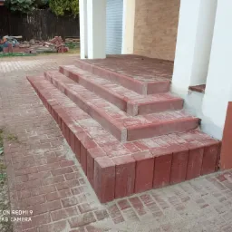 Schody wejściowe wyłożone czerwoną kostką brukową, prowadzące do domu z białymi kolumnami, widoczne pozostałości piasku po fugowaniu.