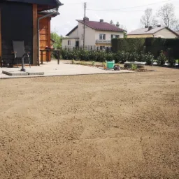 Przygotowanie gruntu pod trawnik z rolki: wyrównana ziemia, rozłożona agrowłóknina, kostka brukowa, budynek gospodarczy w tle, etap prac.