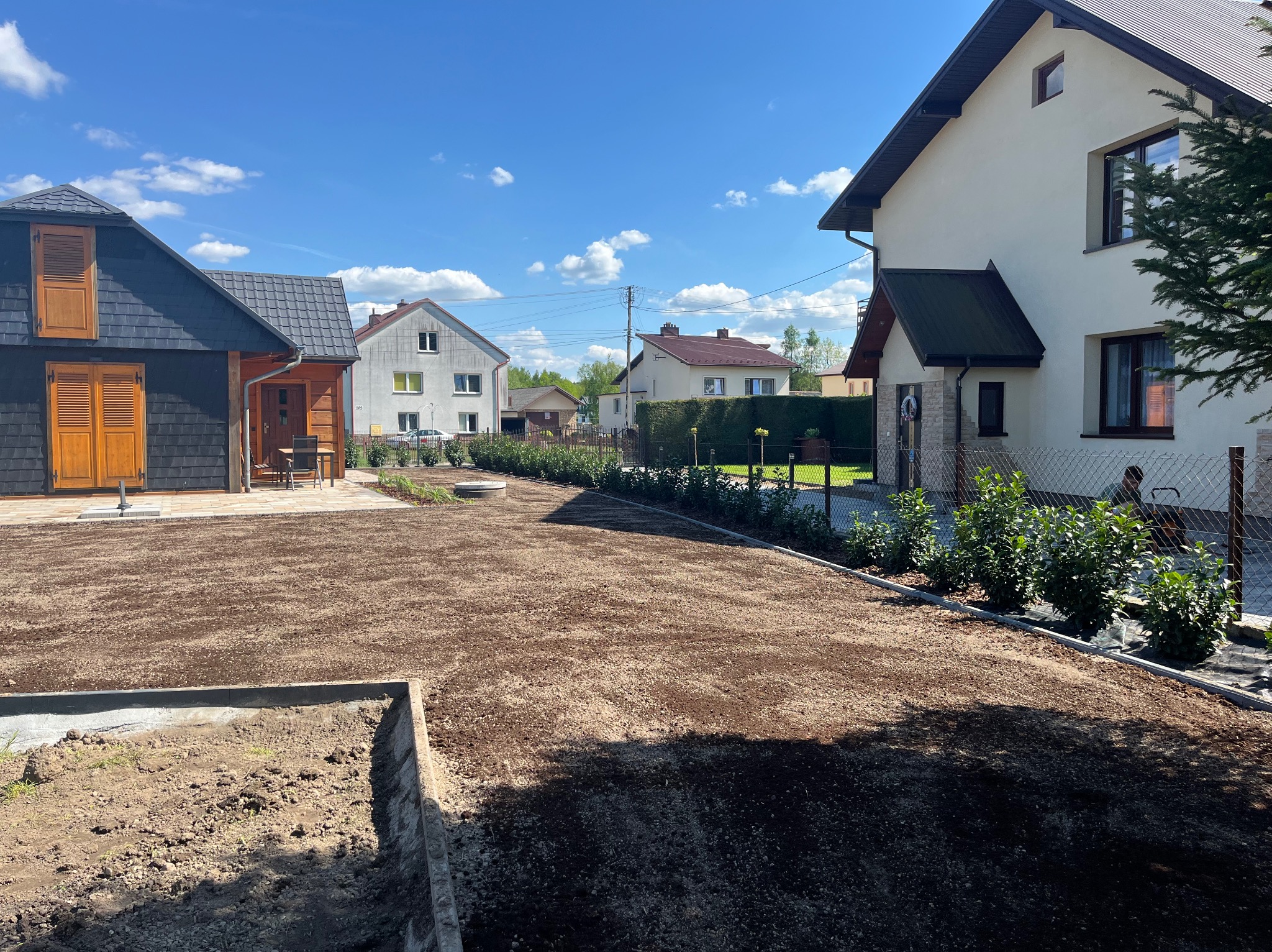 Przygotowany grunt pod trawnik na posesji z dwoma budynkami. Widoczne obrzeża, świeżo posadzona roślinność i osoba w tle. Błękitne niebo z chmurami.