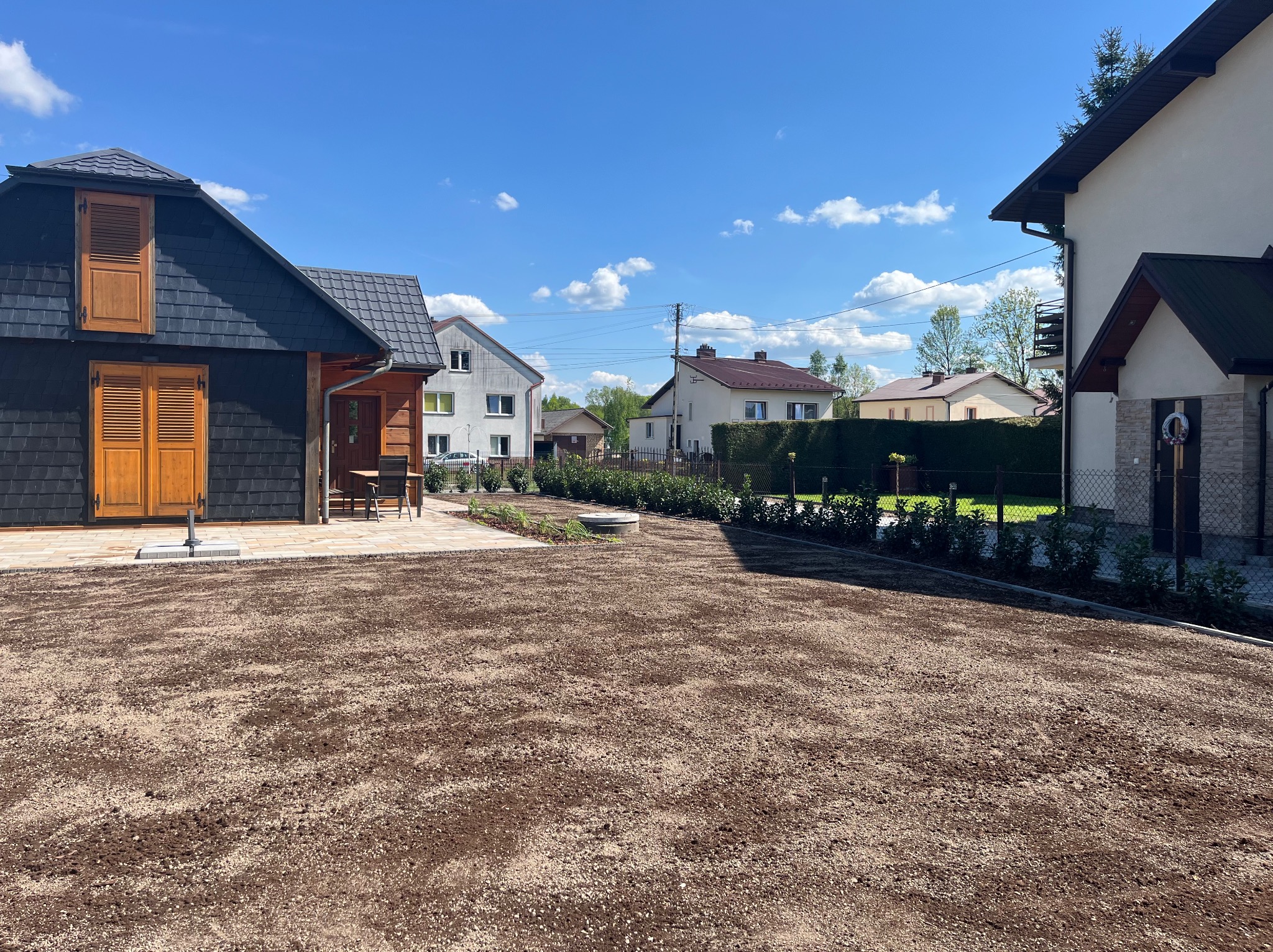 Przygotowany teren pod trawnik z widocznymi obrzeżami, rabatami z niską zielenią i zabudową jednorodzinną w tle, w słoneczny dzień.