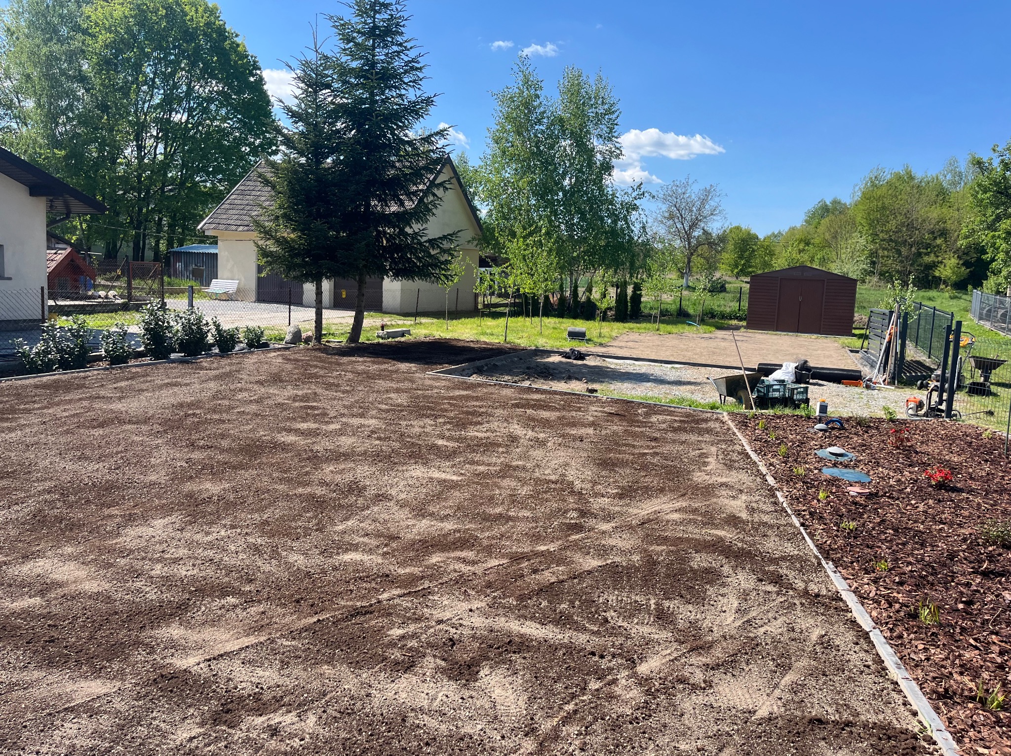 Przygotowany teren pod trawnik z widocznymi śladami po pracy glebogryzarki, obrzeżami z kostki i korą. W tle dom i drzewa pod błękitnym niebem.