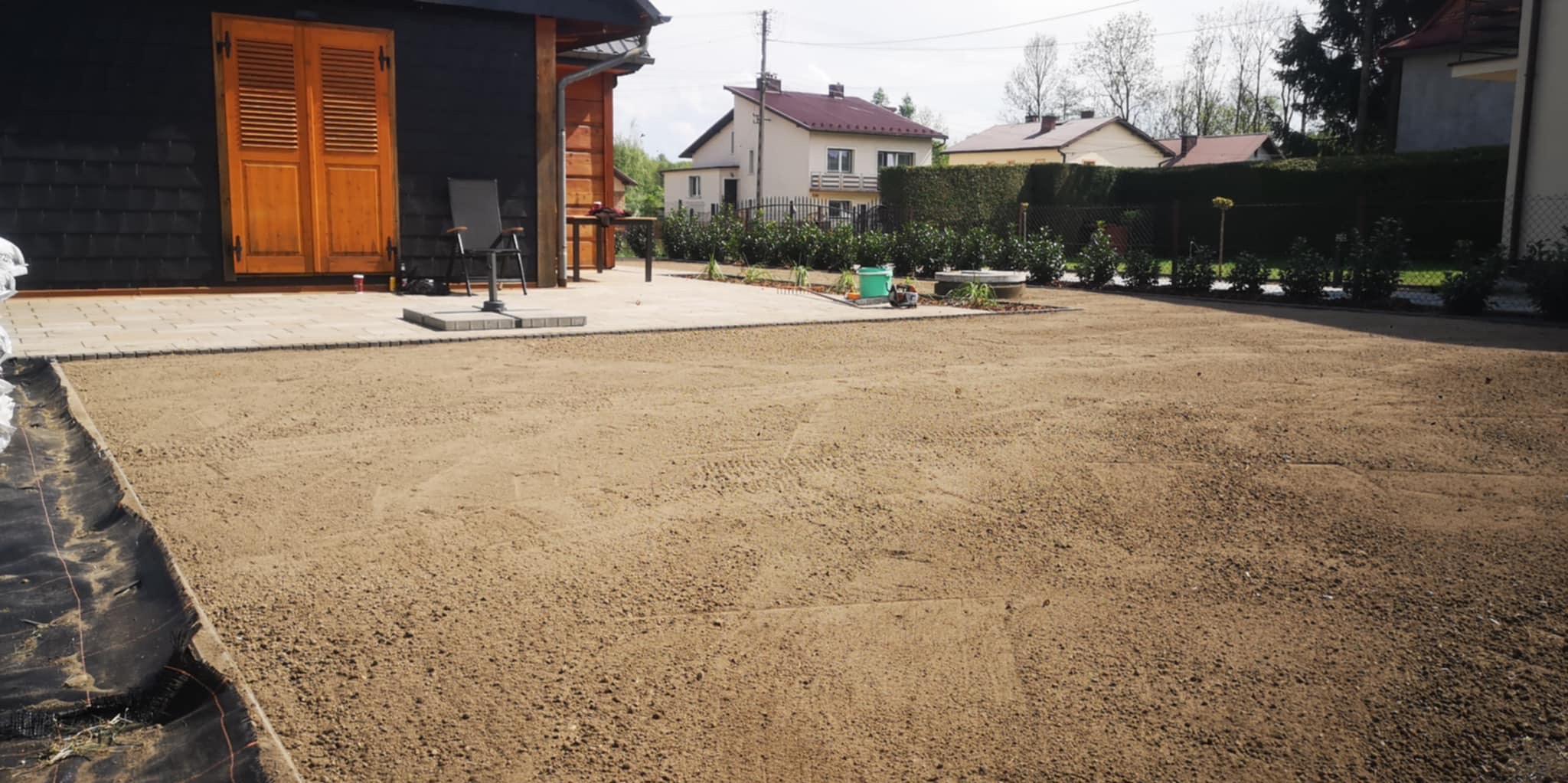 Przygotowanie gruntu pod trawnik z rolki: wyrównana ziemia, rozłożona agrowłóknina, kostka brukowa, budynek gospodarczy w tle, etap prac.