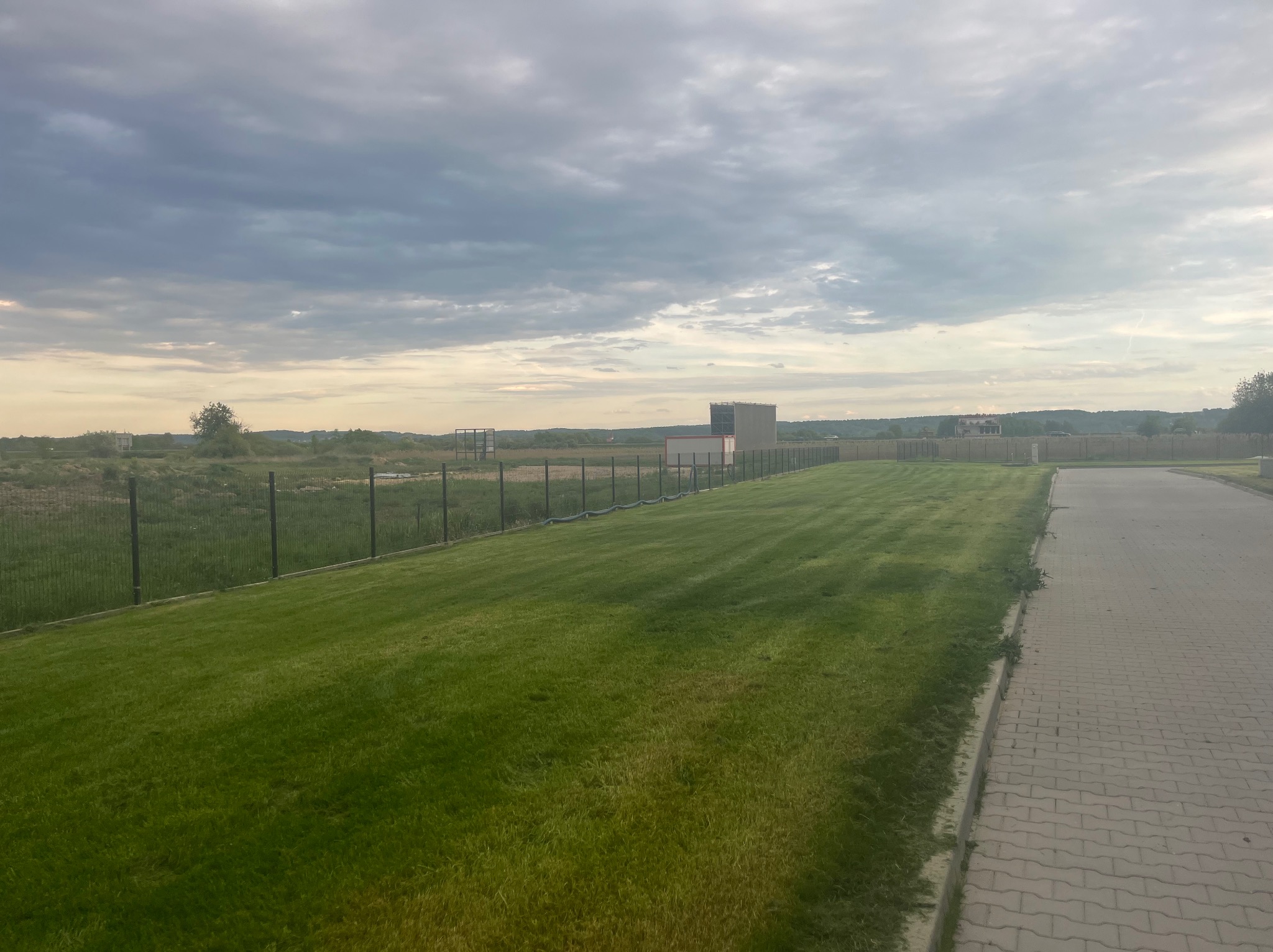 Równomiernie skoszony trawnik przylegający do chodnika z kostki, ogrodzony siatką, pod pochmurnym niebem. Widoczne pasy po koszeniu.