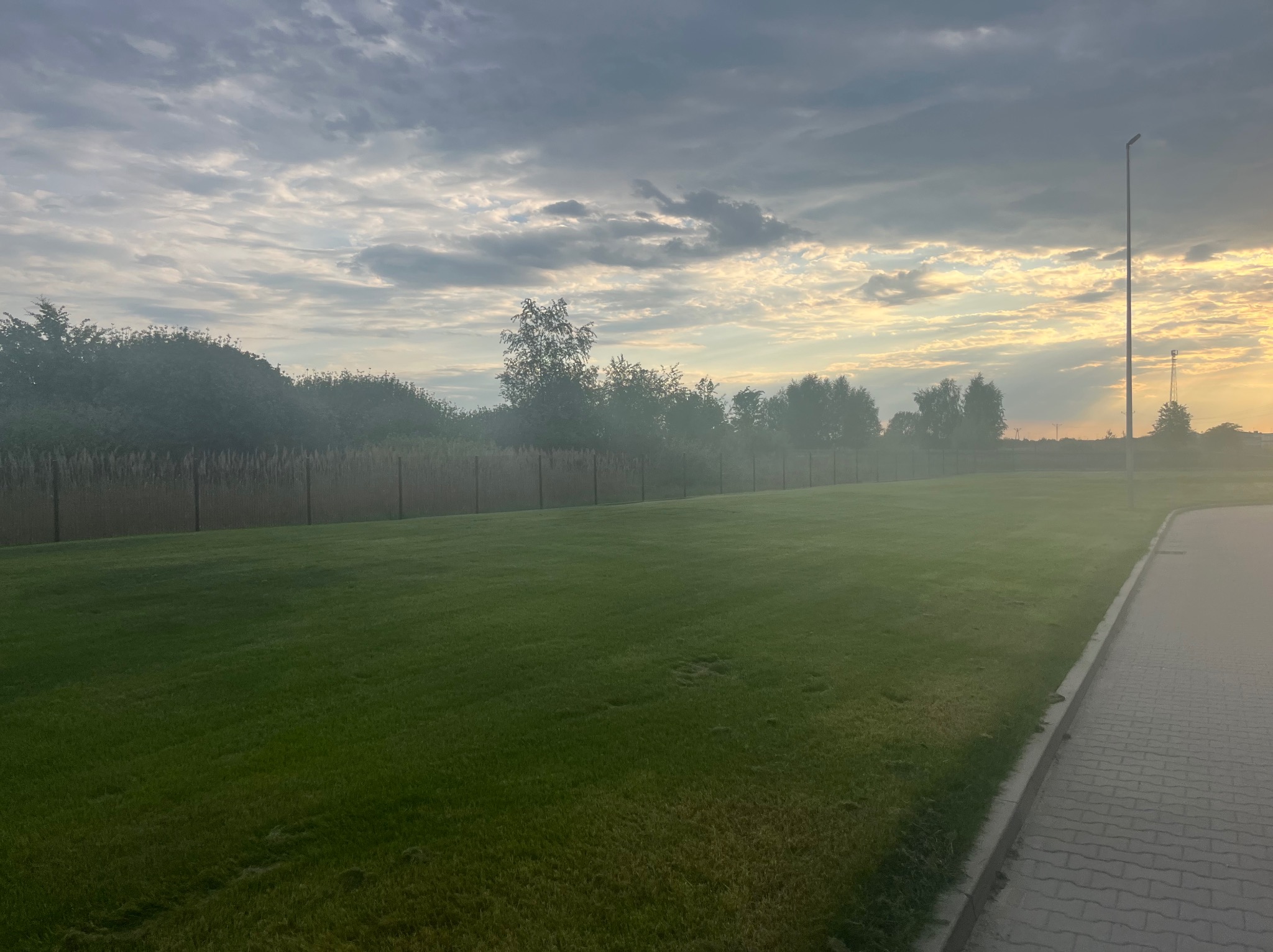 Rozległy trawnik z równo skoszoną trawą, otoczony drzewami i ogrodzeniem, pod pochmurnym niebem o wschodzie słońca. Widoczny chodnik z kostki brukowej i wysoki słup oświetleniowy.