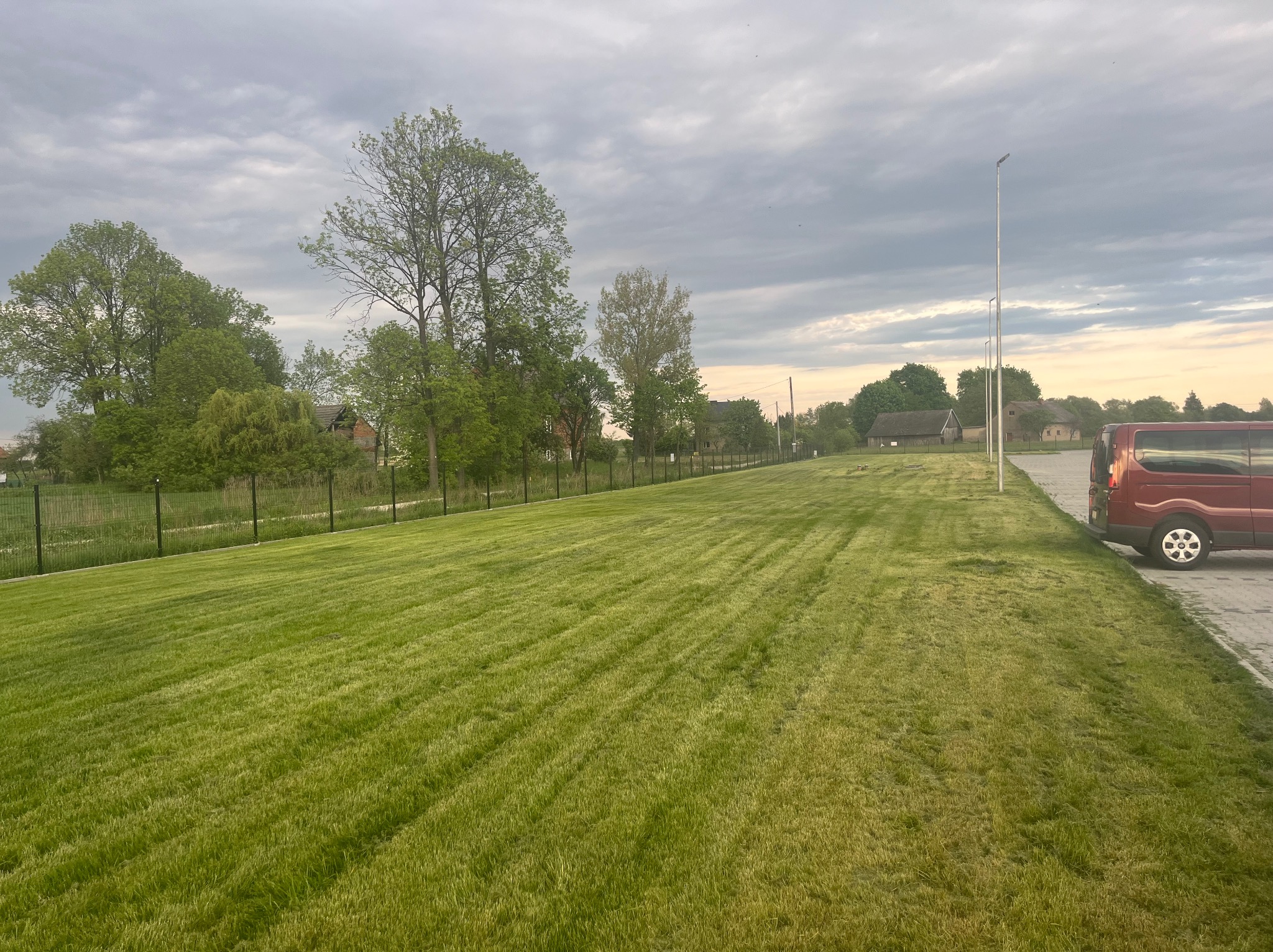 Rozległy trawnik po koszeniu, widoczne ślady po kosiarce, ogrodzenie z siatki, drzewa i budynki w tle, pochmurne niebo. Czerwony samochód zaparkowany obok.