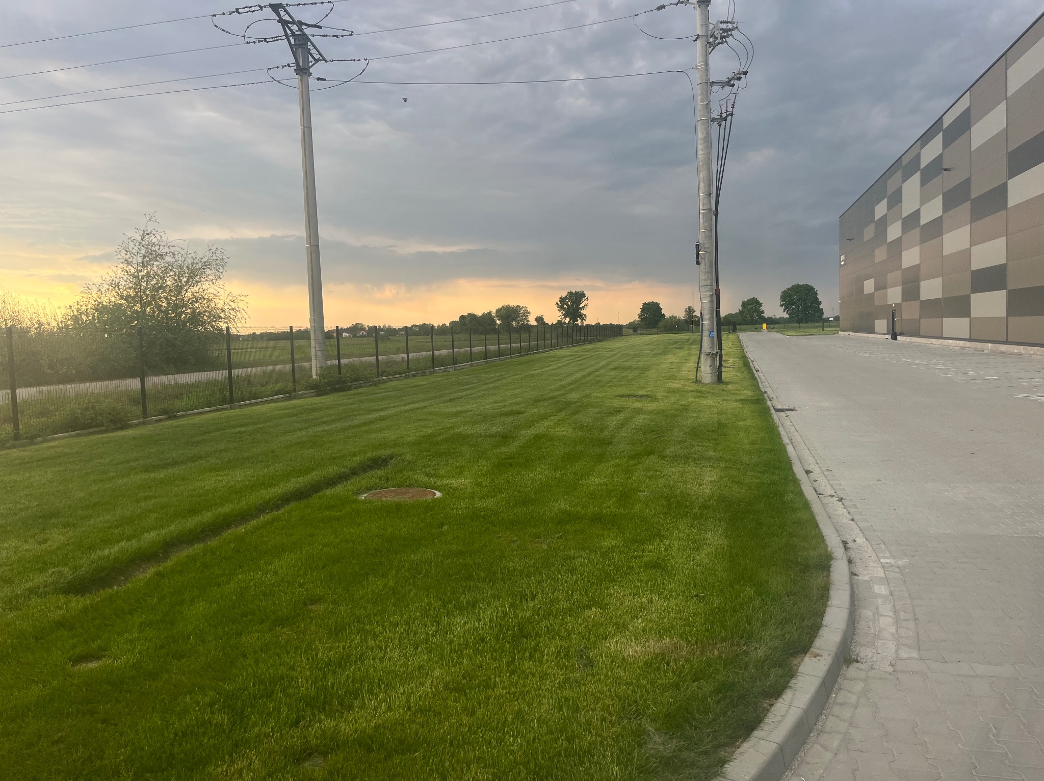 Równomiernie skoszona trawa przy budynku przemysłowym, widok z poziomu gruntu. Słupy energetyczne i ogrodzenie wzdłuż trawnika, zachodzące słońce na horyzoncie.