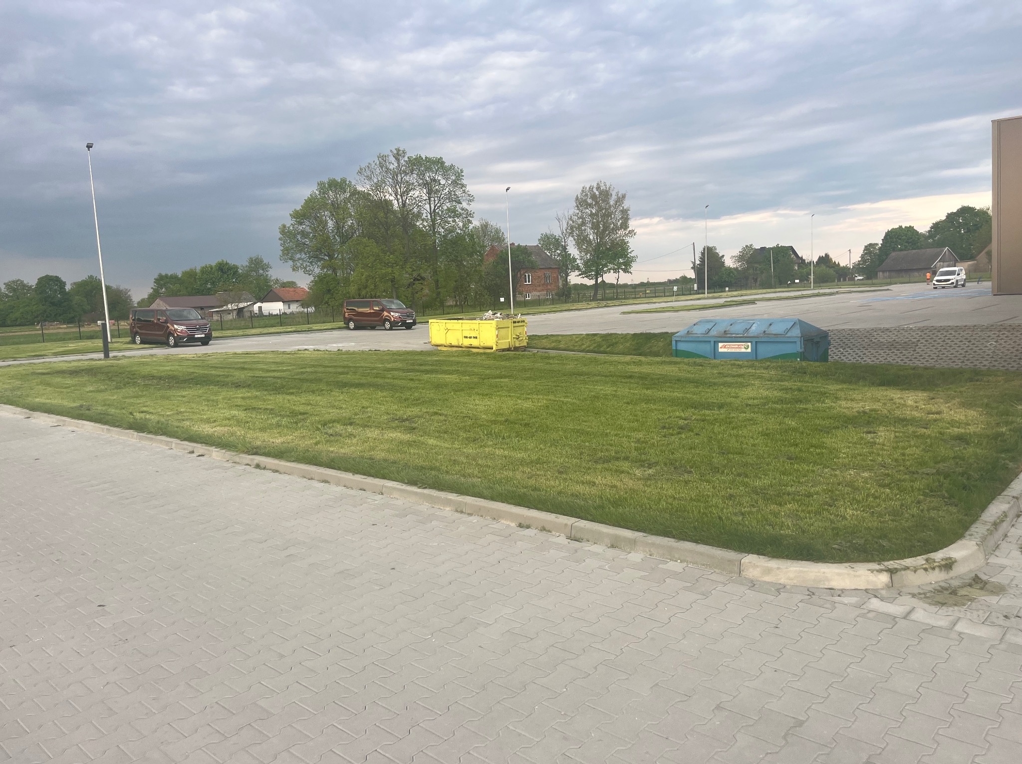 Równy trawnik na posesji z pojazdami i kontenerami na tle pochmurnego nieba. Widok z poziomu chodnika z kostki brukowej.