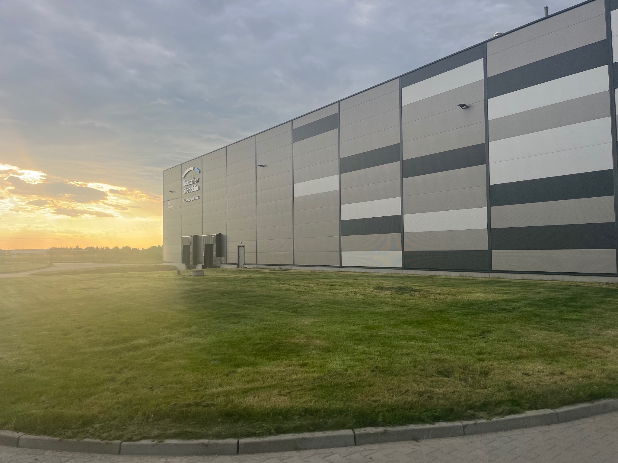 Rozległy trawnik przed nowoczesnym budynkiem przemysłowym o szaro-białej elewacji w porannym świetle. Widoczne ślady koszenia i delikatna mgła nad łąką w tle.