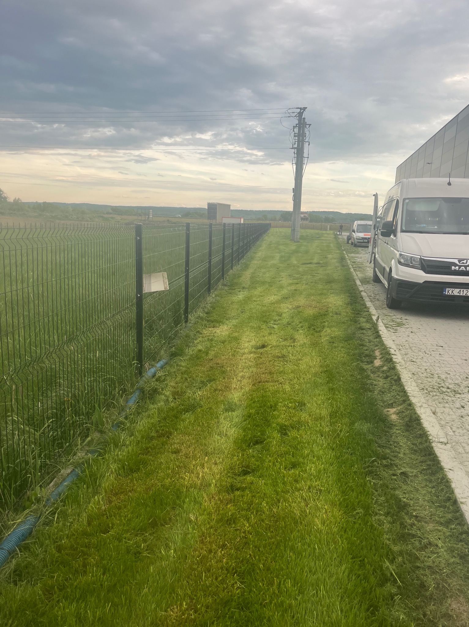 Świeżo skoszona trawa wzdłuż metalowego ogrodzenia i chodnika, z zaparkowanym busem. Widoczne źdźbła trawy po koszeniu. Pochmurne niebo w tle.
