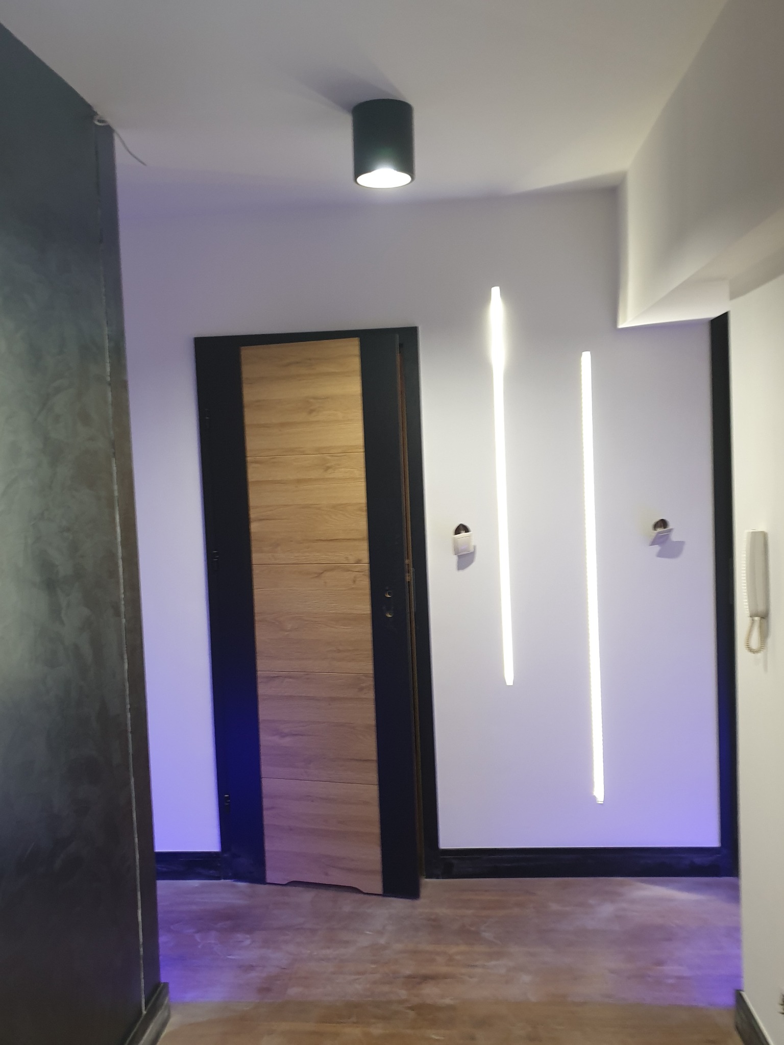 Nowoczesne wnętrze: drewniane drzwi z czarną ramą, białe ściany z pionowymi lampami LED, szary filar i podłoga w odcieniach beżu.