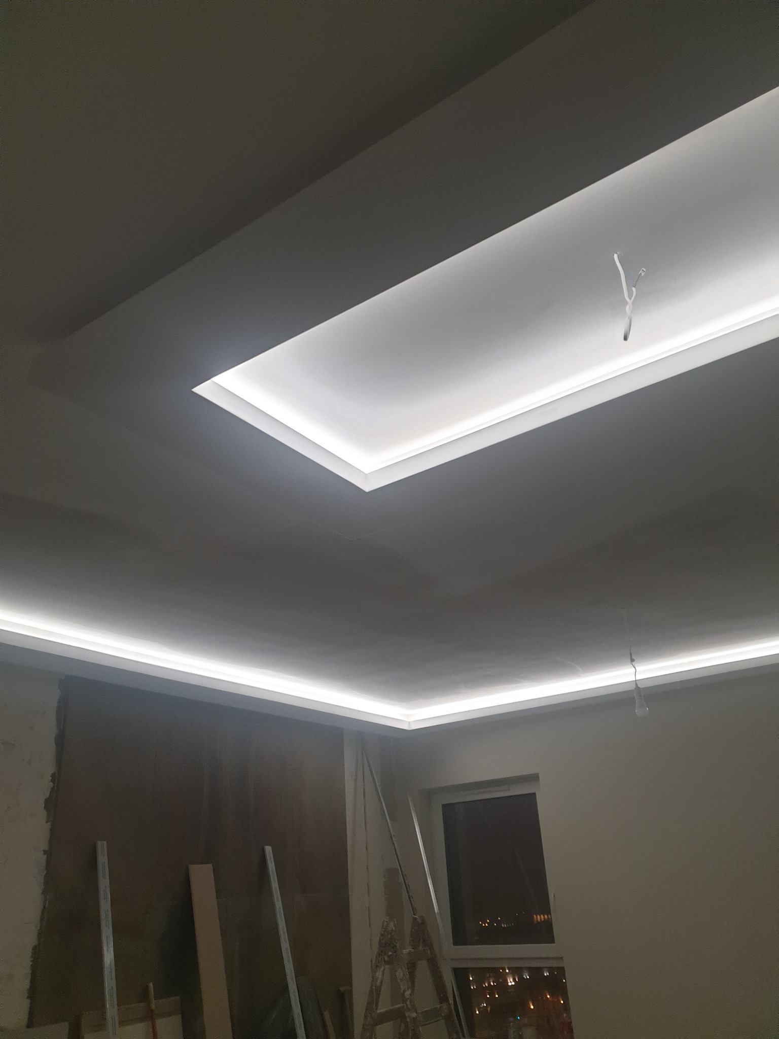 Wnętrze w trakcie prac wykończeniowych: sufit z podwieszaną konstrukcją i oświetleniem LED, ściana przygotowana do dalszych prac, widoczna drabina i materiały budowlane.