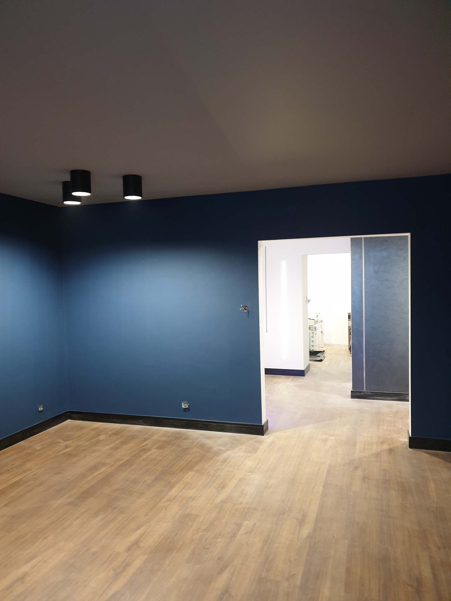 Wnętrze po remoncie: ściany pomalowane na ciemny niebieski kolor, drewniana podłoga, czarne listwy przypodłogowe, sufit w kolorze szarym, oświetlenie punktowe, widoczne gniazdka elektryczne...