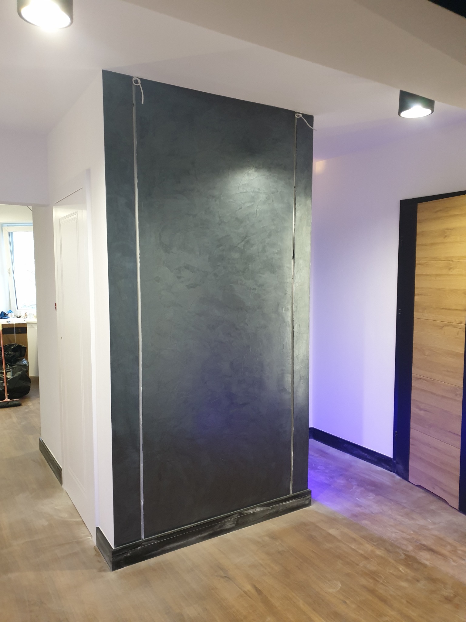 Ściana w stylu loft, wykończona szarą, metalizującą farbą dekoracyjną, z widocznymi pionowymi paskami imitującymi metalowe profile, w nowoczesnym wnętrzu z drewnianą podłogą i drzwiami.