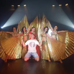 Występ zespołu tanecznego w kostiumach z błyszczącego, złotego materiału, podświetlonych reflektorami na czarnej scenie, z mężczyzną w dżinsowych szortach klęczącym na pierwszym planie.