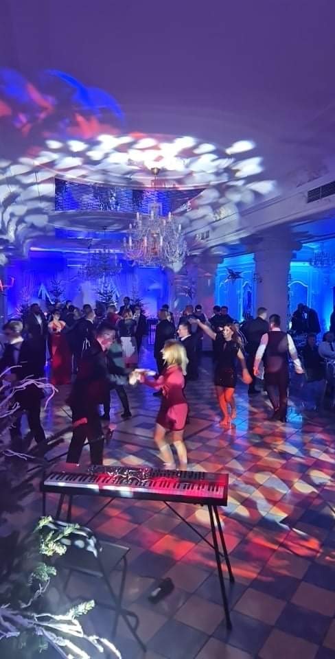 Sala balowa z parkietem w szachownicę, bawiący się ludzie, oświetlenie dyskotekowe i kryształowy żyrandol, na pierwszym planie keyboard na statywie.