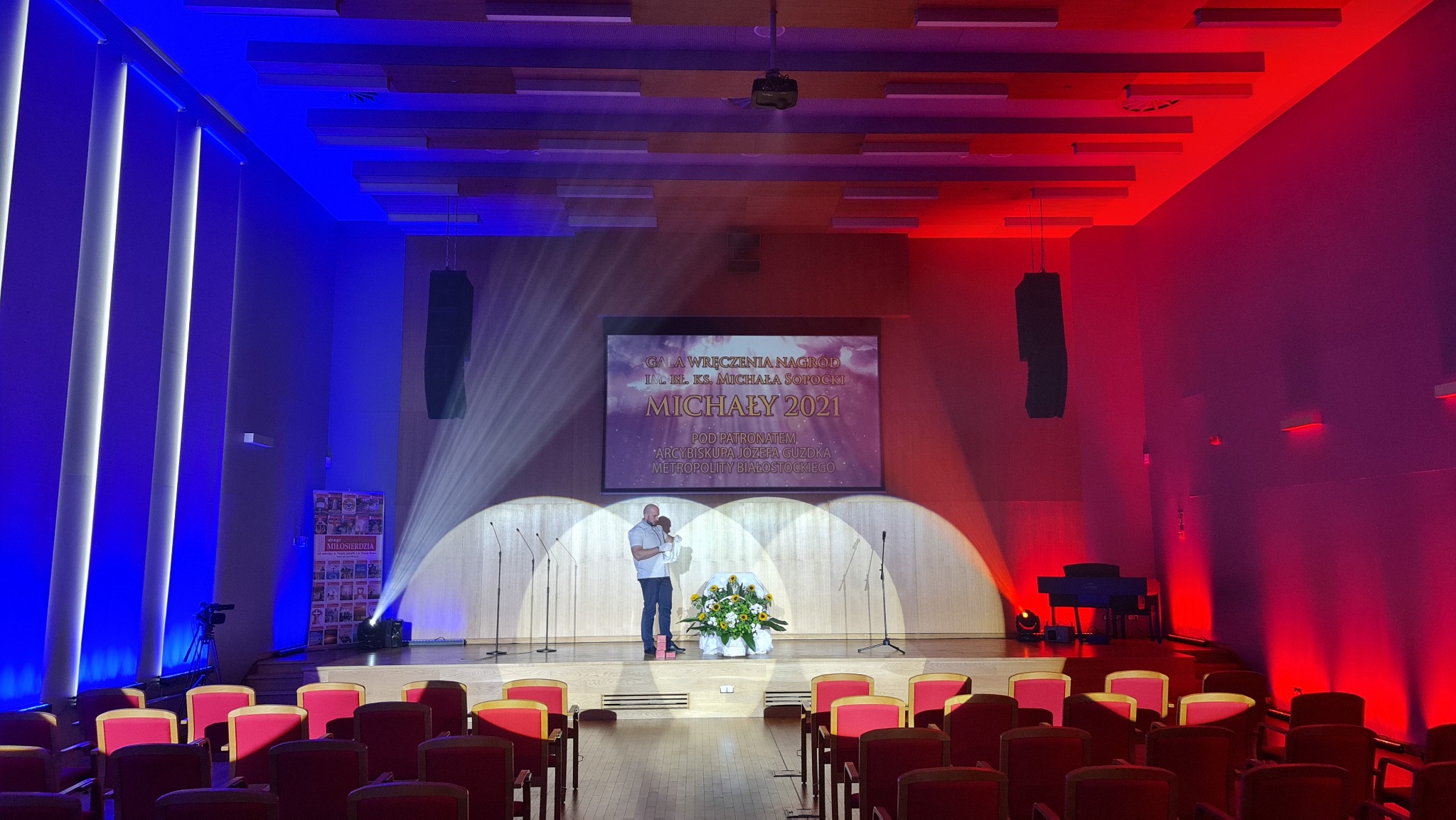 Sala widowiskowa z oświetleniem w kolorach niebieskim i czerwonym, scena z ekranem wyświetlającym napis 'Gala wręczenia nagród Michały 2021', mężczyzna przemawia do mikrofonu, w pierwszym rzędzie...