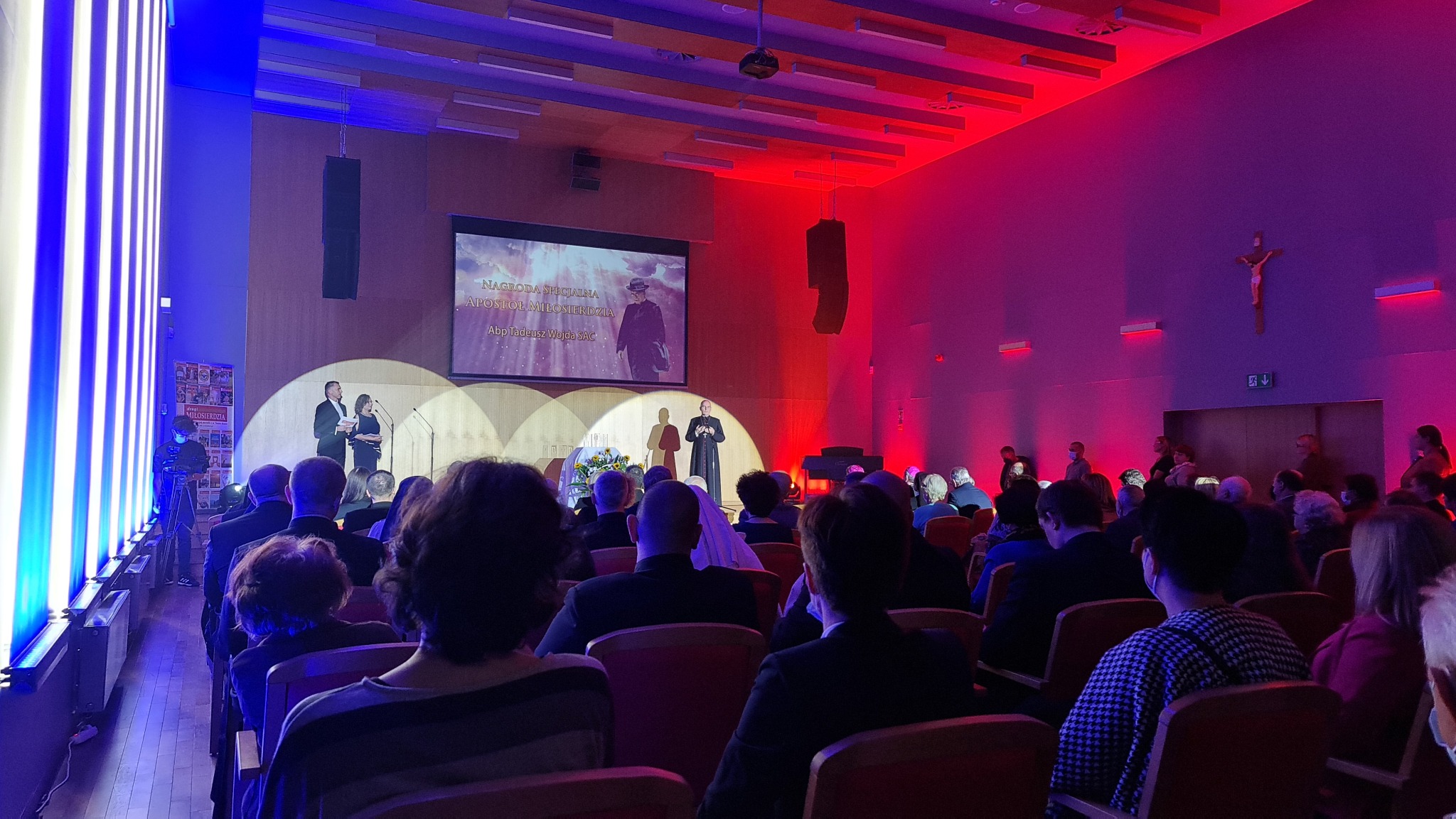 Sala konferencyjna oświetlona na niebiesko i czerwono, z publicznością siedzącą na krzesłach, na scenie prezenterzy i duchowny, ekran z prezentacją w tle.