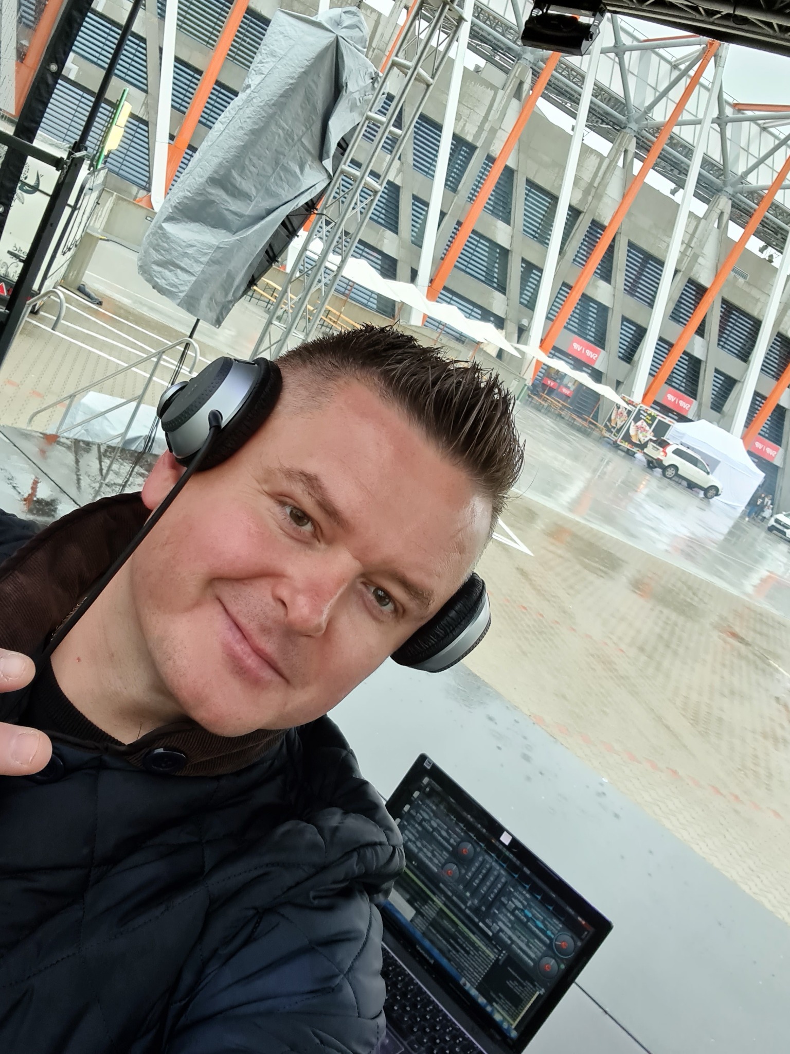 Mężczyzna w słuchawkach, przed laptopem z oprogramowaniem DJ-skim, na tle stadionu podczas deszczowej pogody, widoczne elementy konstrukcji scenicznej.