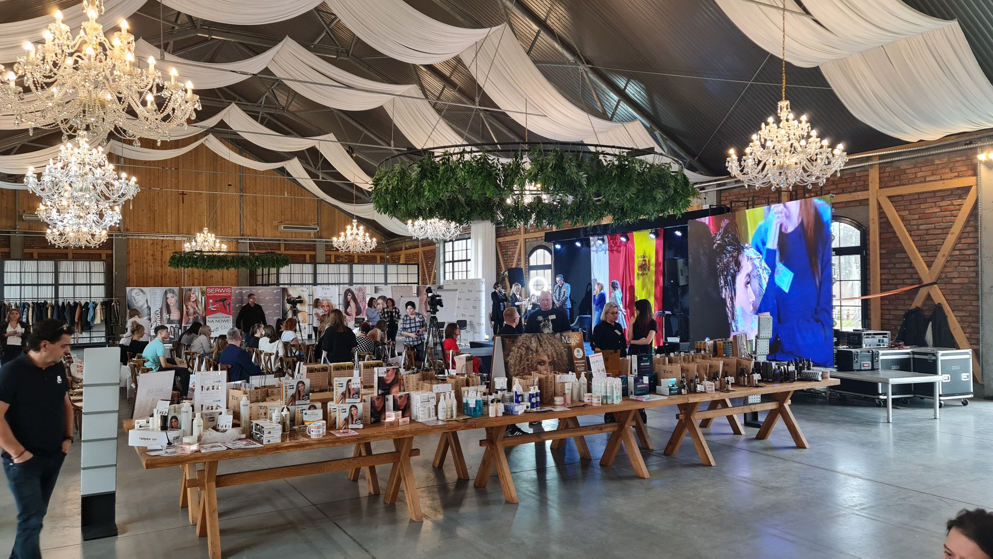 Wnętrze hali z dekoracjami, stołami z produktami kosmetycznymi, sceną w tle i licznymi osobami. Na suficie wiszą żyrandole i białe materiały.