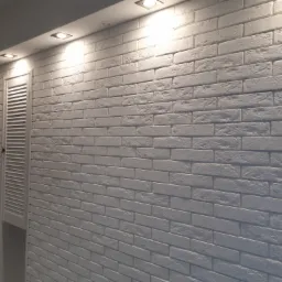 Ściana wykończona białymi cegłami dekoracyjnymi, oświetlona punktowymi lampami sufitowymi, obok widoczne białe drzwi z żaluzjami.