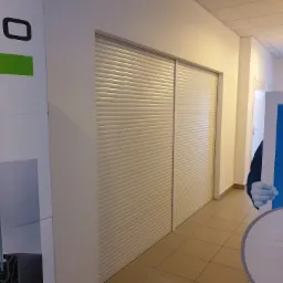 Białe rolety wewnętrzne zwinięte na oknie, widoczne z boku w korytarzu biurowym, obok reklama firmy.