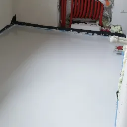 Wylewka samopoziomująca w trakcie realizacji, widoczne rury grzewcze, pianka montażowa przy ścianach, surowy stan wykończenia ścian.