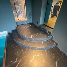 MAX HOME GLAZURNICTWO Szymon Pomarański - Czarne marmurowe płytki z białymi żyłami, ułożone na schodach z mozaiką, prowadzących do basenu. Widok na siłownię w tle, przez otwarte drzwi.