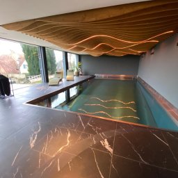 MAX HOME GLAZURNICTWO Szymon Pomarański - Nowoczesny kryty basen z marmurową posadzką i drewnianym sufitem z podświetleniem LED. Leżaki przy oknie z widokiem na zieleń. Minimalistyczny design.