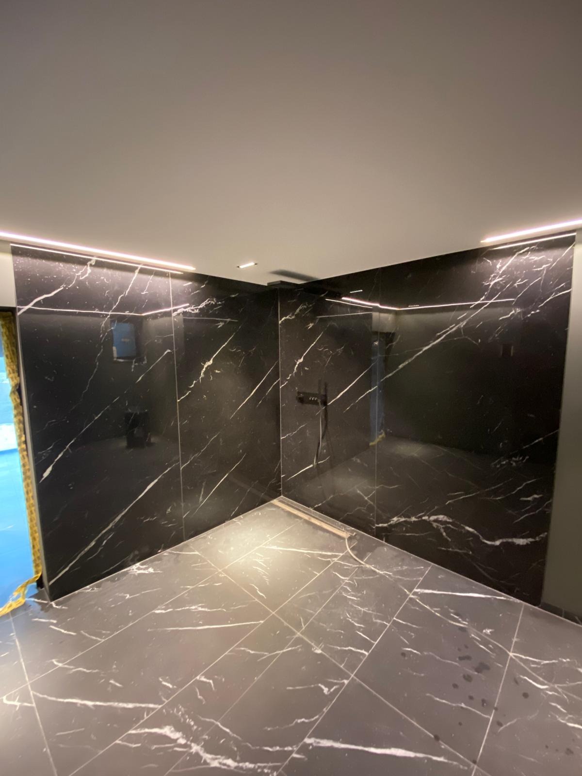 Wykończona kabina prysznicowa z czarnymi płytkami imitującymi marmur, podświetlona listwami LED. Widoczny odpływ liniowy i bateria prysznicowa.