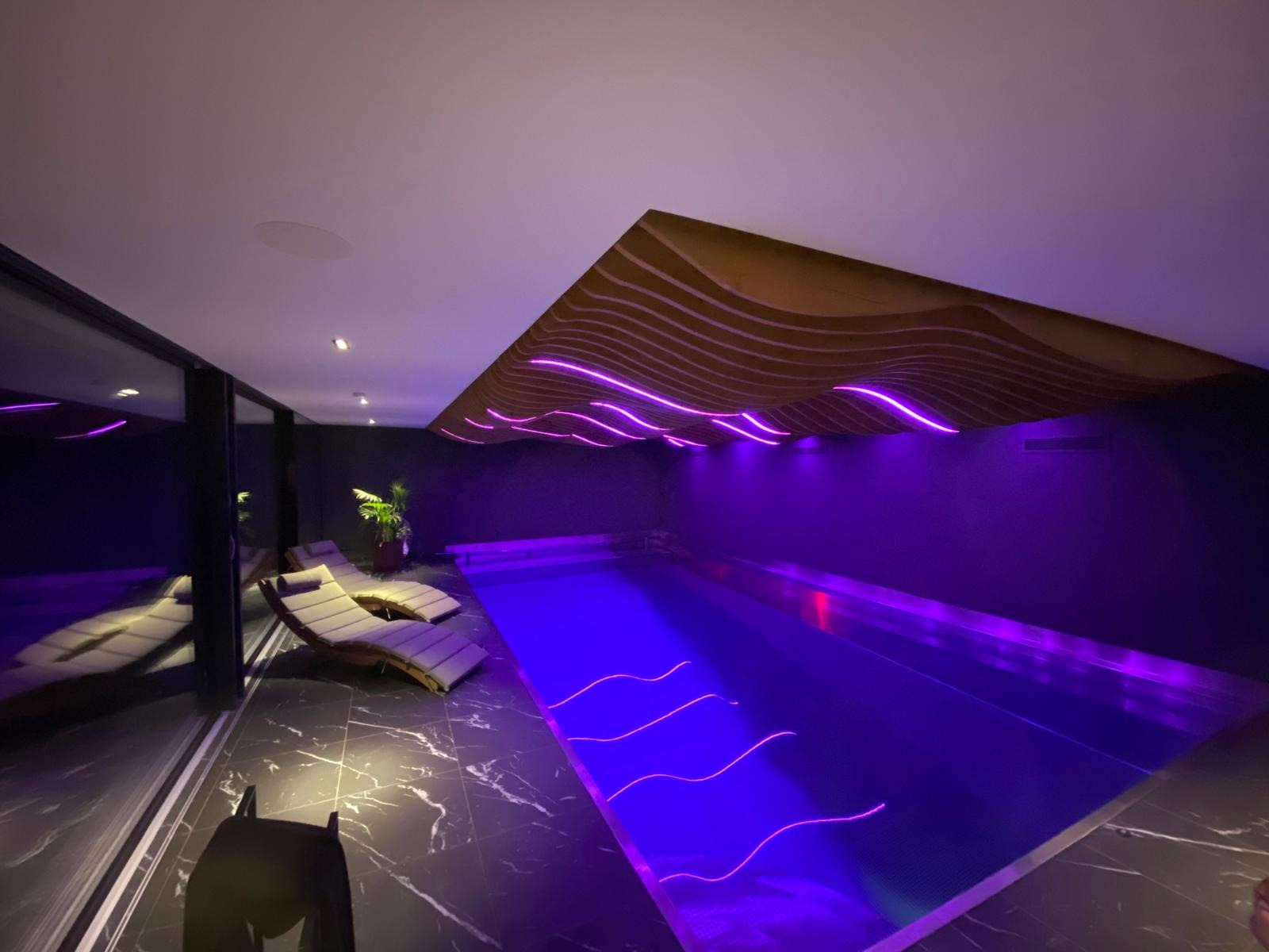 Nowoczesny kryty basen z marmurową posadzką, leżakami i podwieszanym sufitem z oświetleniem LED. Minimalistyczny design i relaksująca atmosfera.
