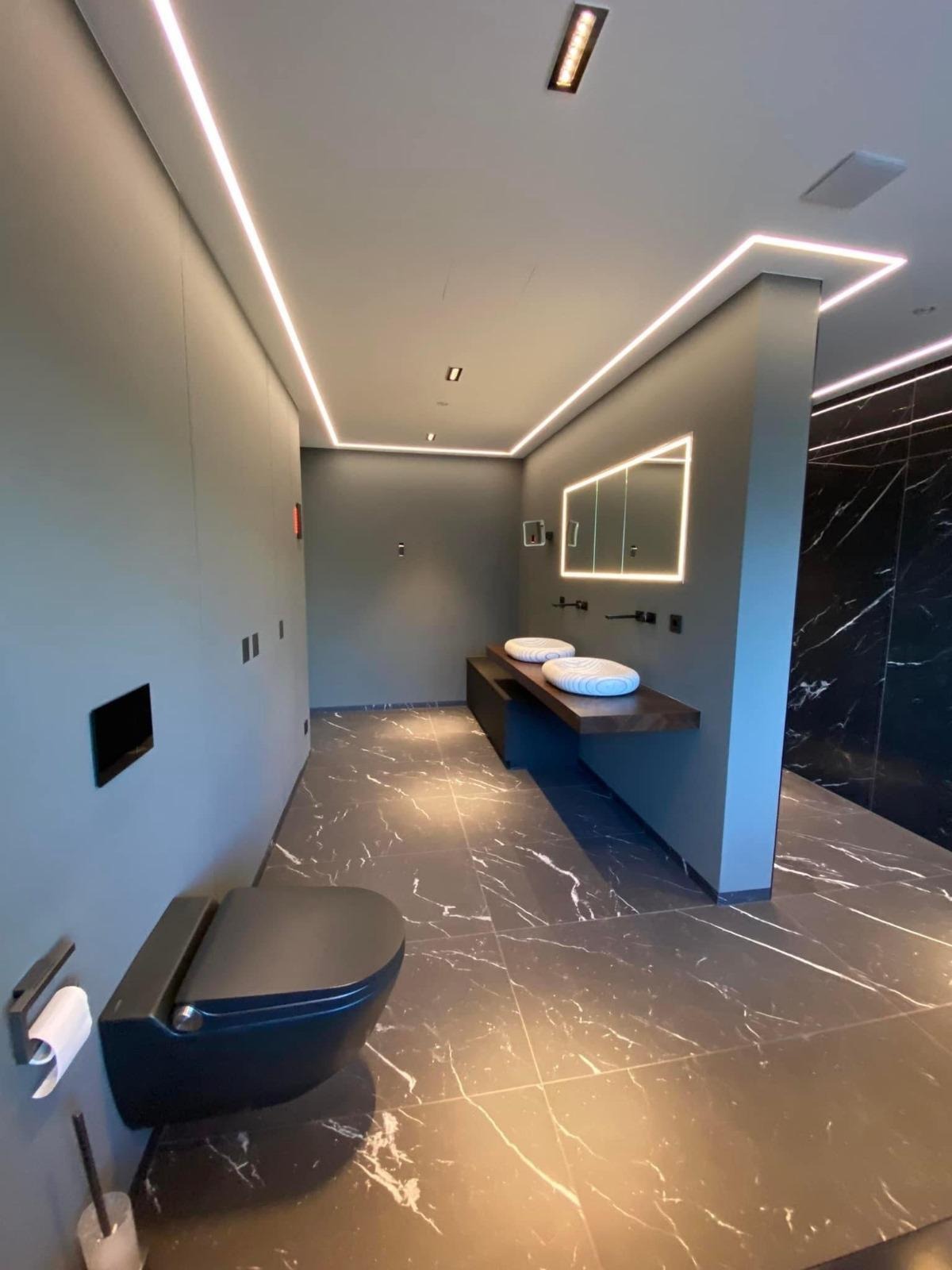 Nowoczesna łazienka z szarą armaturą, marmurową podłogą i podświetlanym lustrem. Uwagę przykuwają minimalistyczne umywalki i oświetlenie LED wzdłuż sufitu.