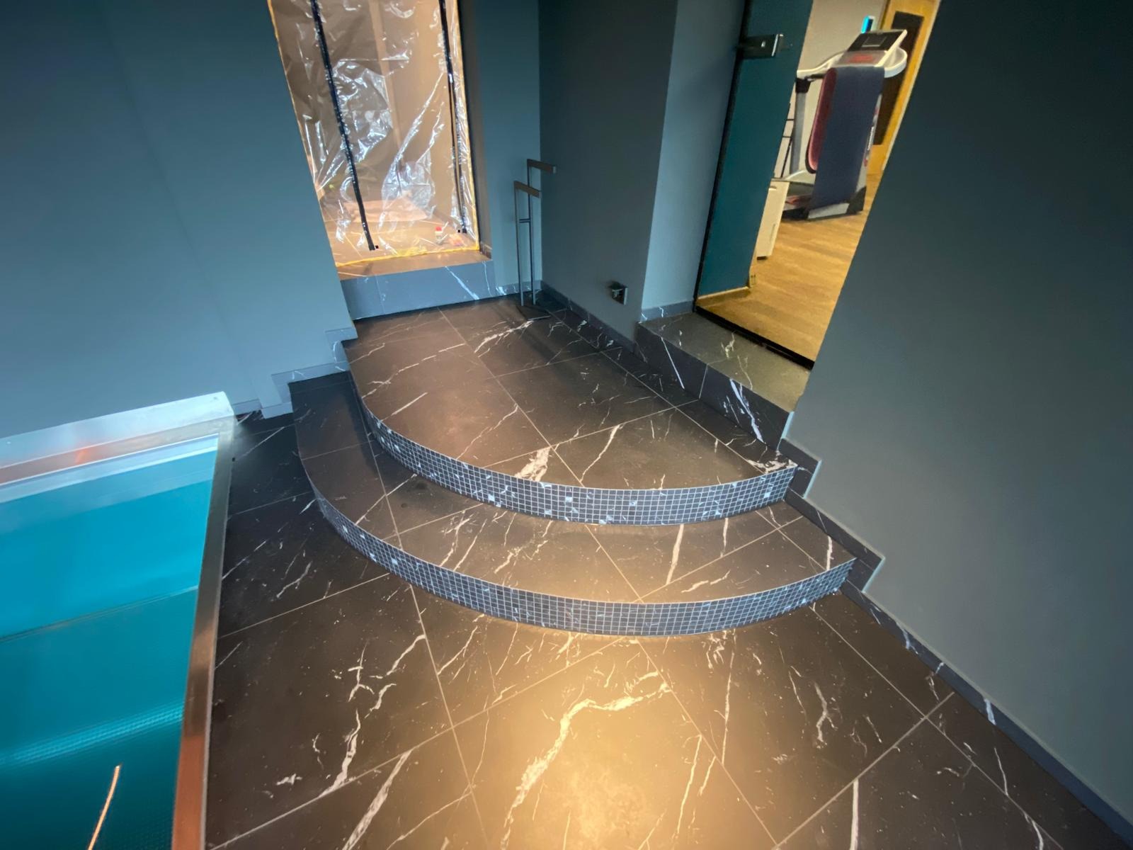 Czarne marmurowe płytki z białymi żyłami, ułożone na schodach z mozaiką, prowadzących do basenu. Widok na siłownię w tle, przez otwarte drzwi.