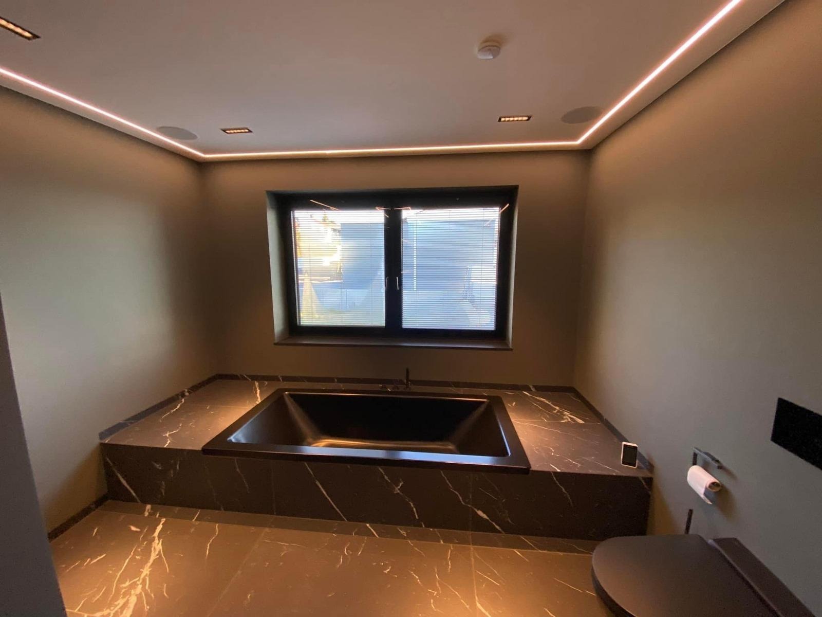 Nowoczesna łazienka z czarną wanną obudowaną marmurem, podświetleniem LED i marmurową podłogą. Minimalistyczny design, okno z żaluzjami.