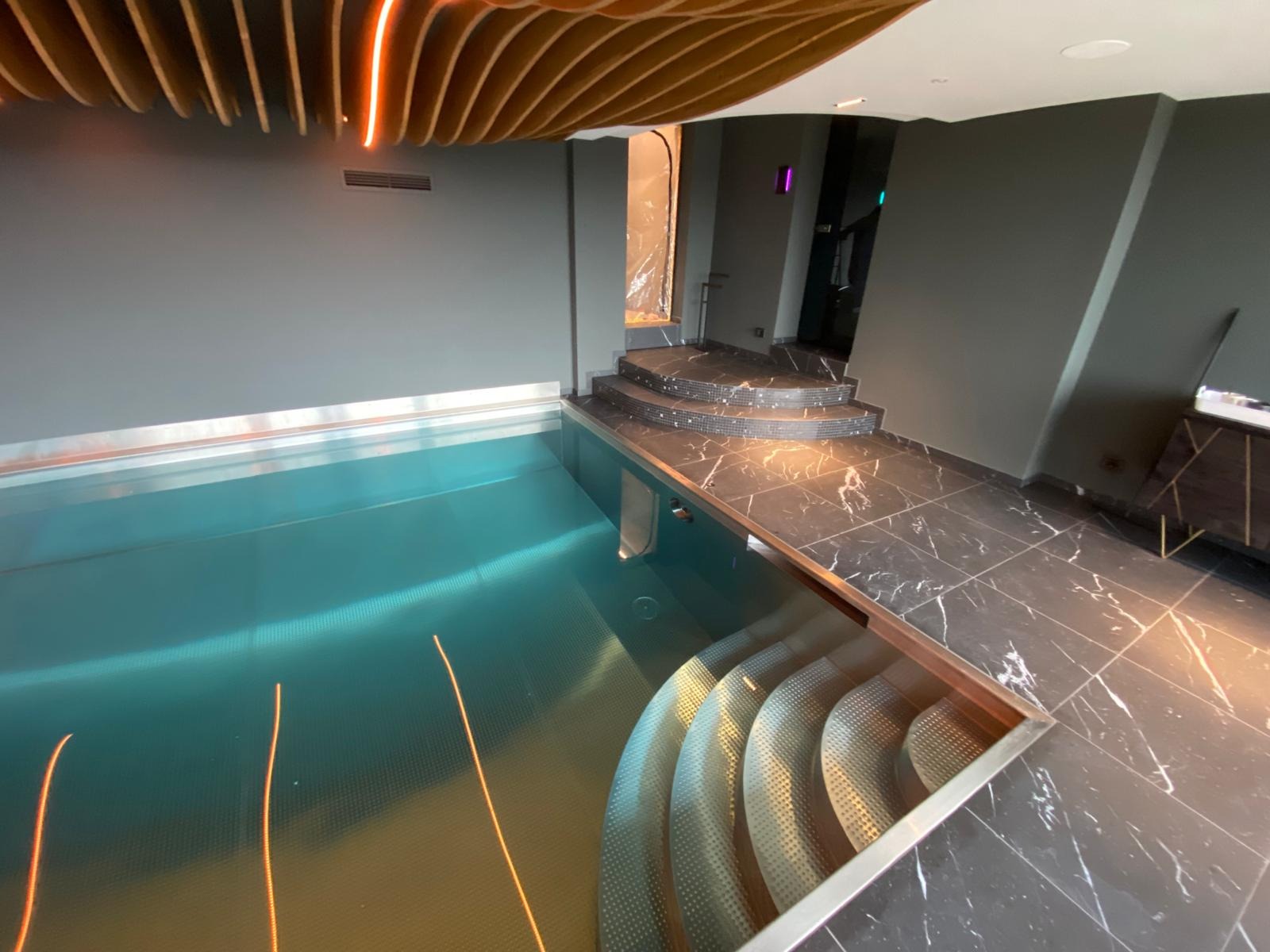 Nowoczesny basen w pomieszczeniu z marmurową posadzką i schodami. Oświetlenie LED podkreśla drewniany sufit i turkusową wodę. Minimalistyczny design.