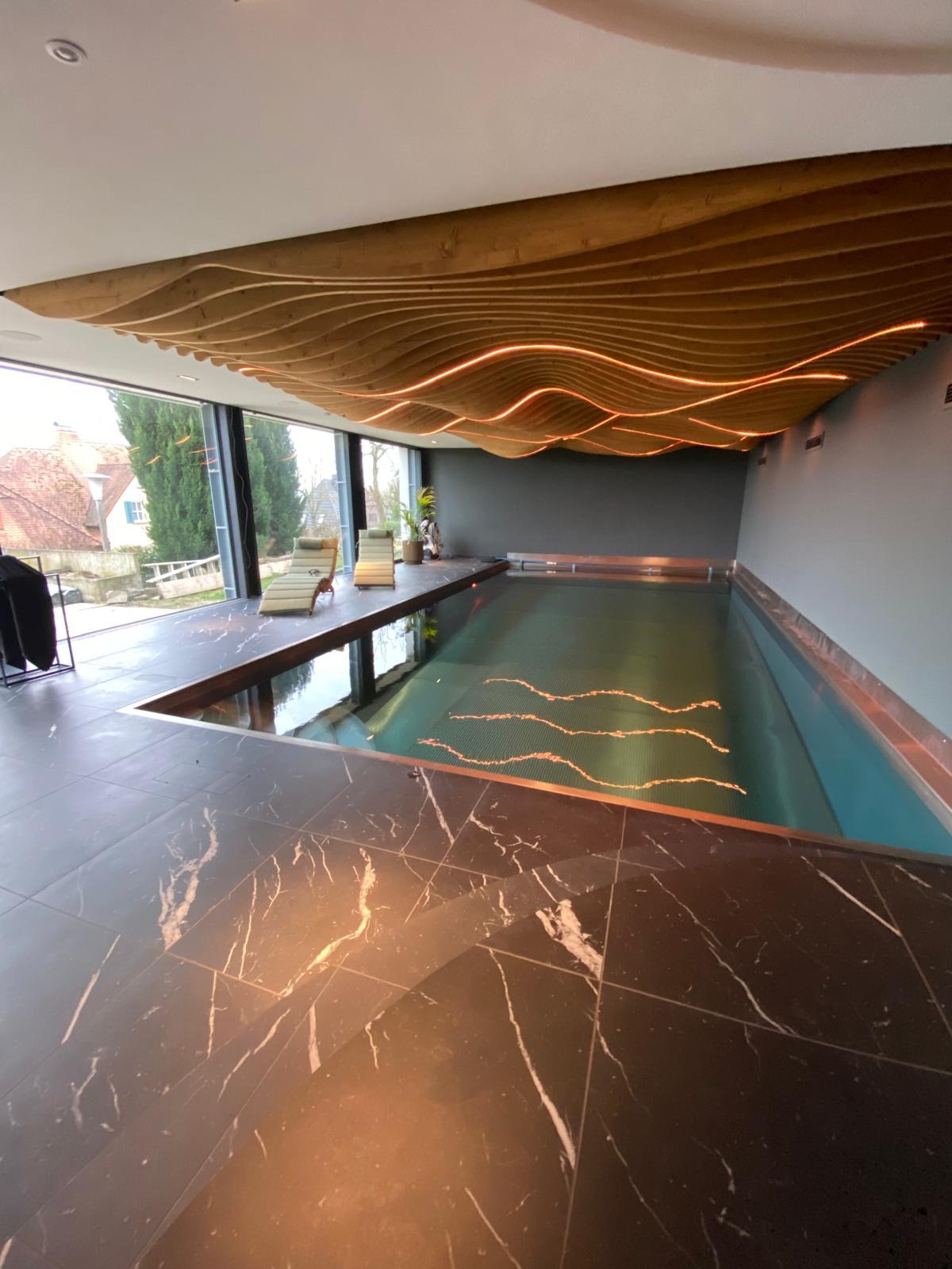 Nowoczesny kryty basen z marmurową posadzką i drewnianym sufitem z podświetleniem LED. Leżaki przy oknie z widokiem na zieleń. Minimalistyczny design.