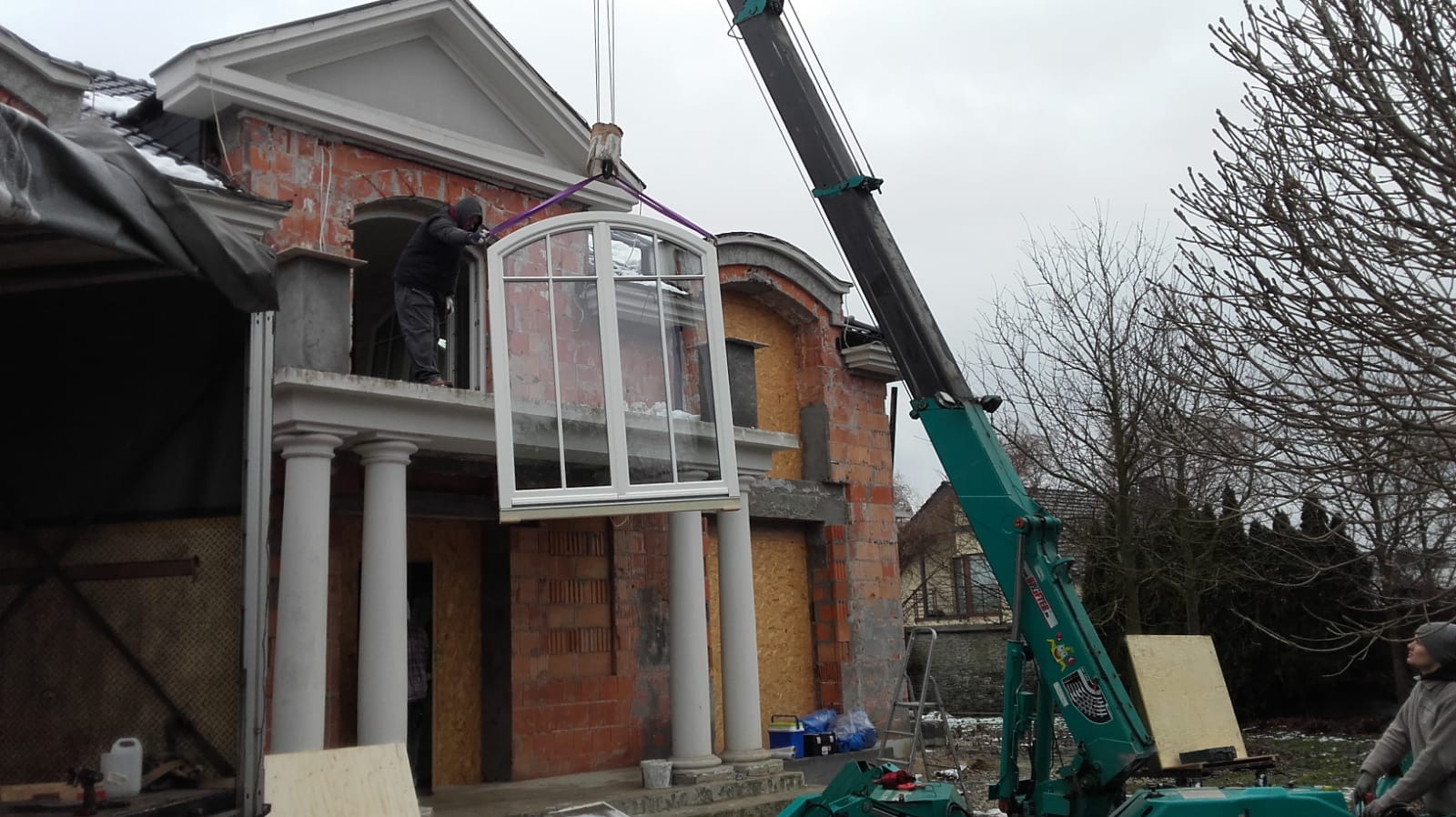 Montaż okna o łukowym kształcie, podnoszonego przez dźwig na tle surowej elewacji budynku w trakcie budowy, widoczny pracownik na balkonie.