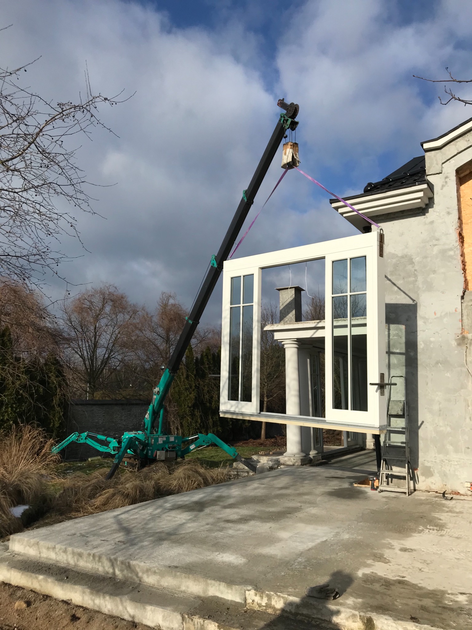 Montaż okna panoramicznego przy użyciu dźwigu teleskopowego na tle częściowo wykończonego budynku w słoneczny dzień.