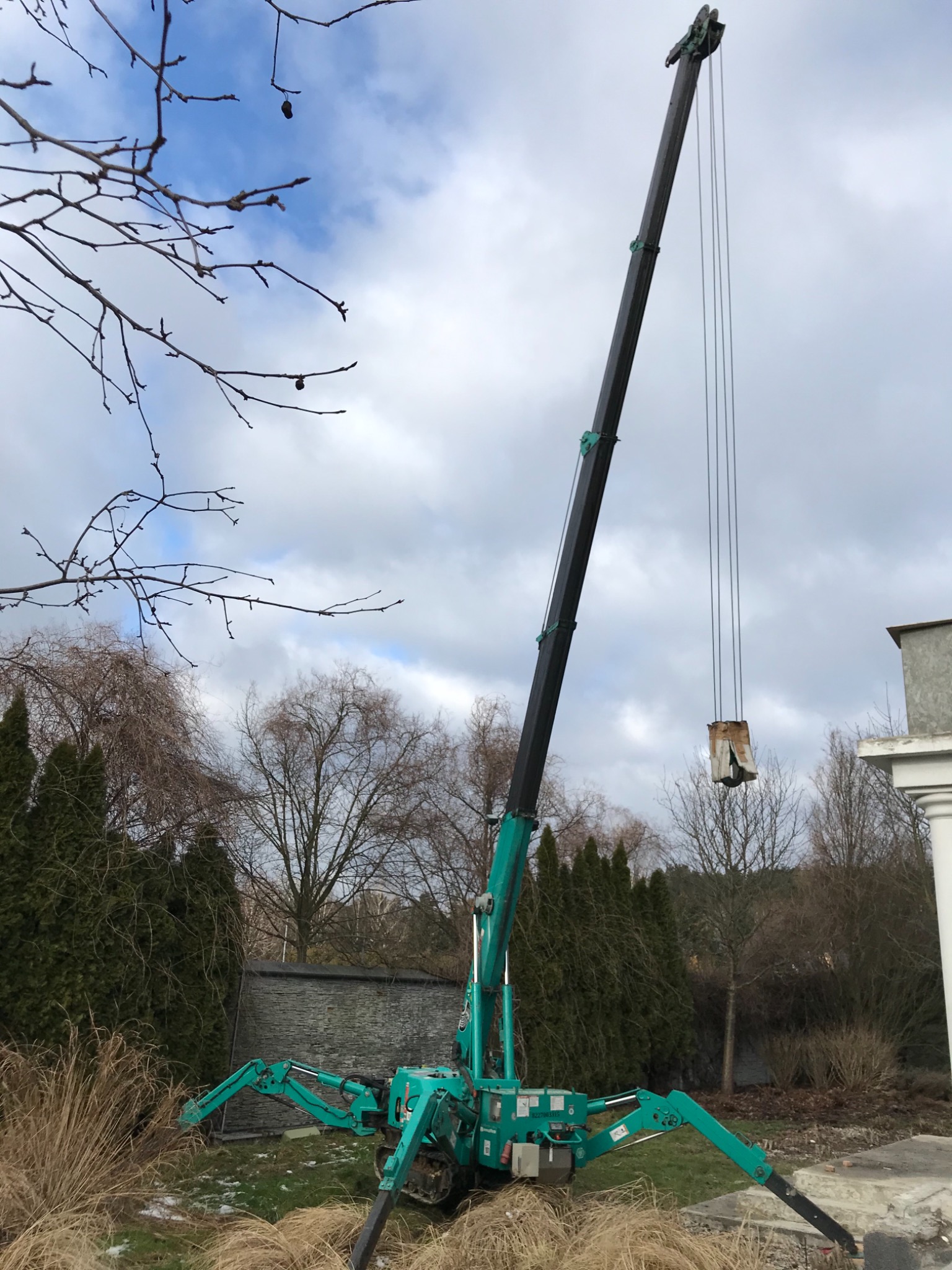 Zielony mini-dźwig pająk z wysuniętym ramieniem podnosi mały, brązowy ładunek na tle pochmurnego nieba i drzew bez liści.