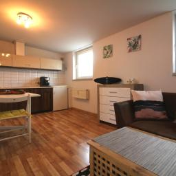 SILFOX nieruchomości Sebastian Lis - Jasne studio z aneksem kuchennym, drewnianą podłogą, brązową kanapą z dekoracyjnymi poduszkami i stolikiem kawowym z szarą podkładką.