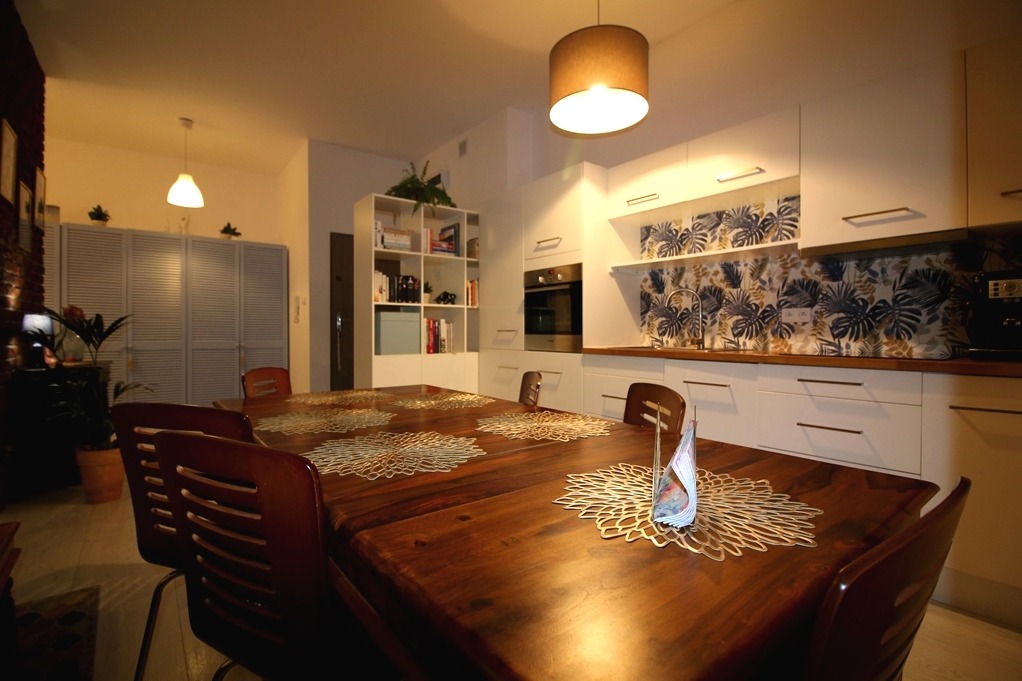 Jasne wnętrze kuchni z drewnianym stołem, na którym leżą ażurowe podkładki, w tle białe szafki z motywem liści oraz regał z książkami.