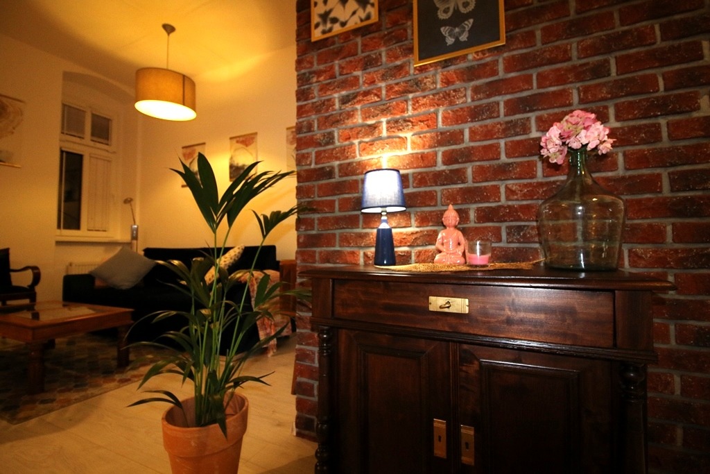 Ciepłe, przytulne wnętrze z ceglaną ścianą, drewnianą komodą z dekoracjami (lampa, figurka Buddy, kwiaty w wazonie) i rośliną doniczkową w terakotowej doniczce, widok z salonu z sofą i stolikiem...