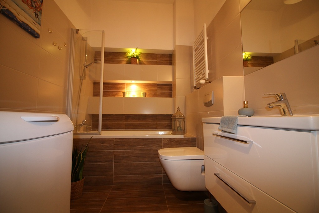 Nowoczesna łazienka z wanną obudowaną brązowymi płytkami, podświetlanymi wnękami, białą umywalką z chromowaną baterią i pralką z boku.