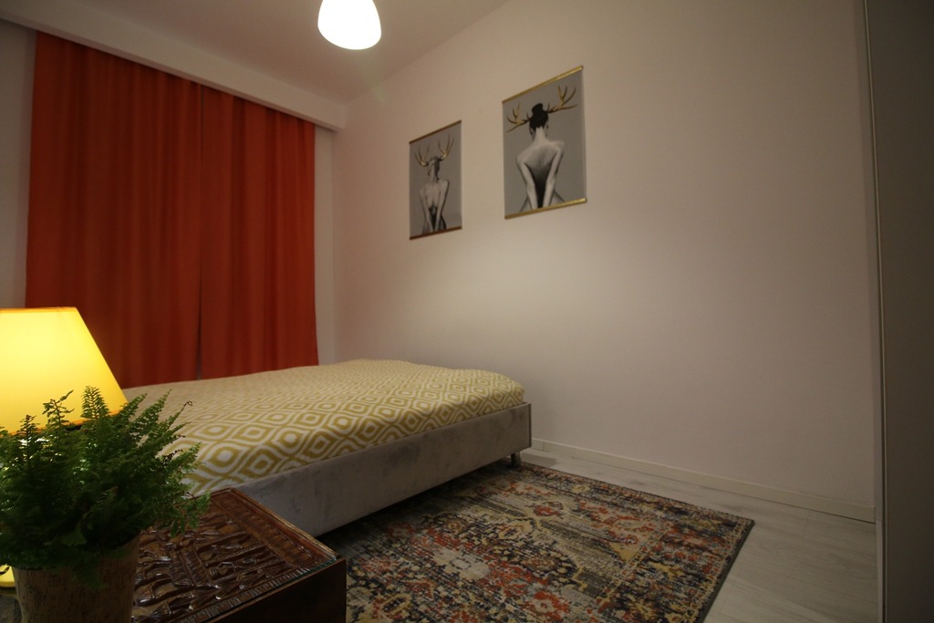 Pokój z łóżkiem, pomarańczowymi zasłonami, dywanem w stylu vintage i dwoma obrazami z motywem kobiety z porożem jelenia na ścianie.