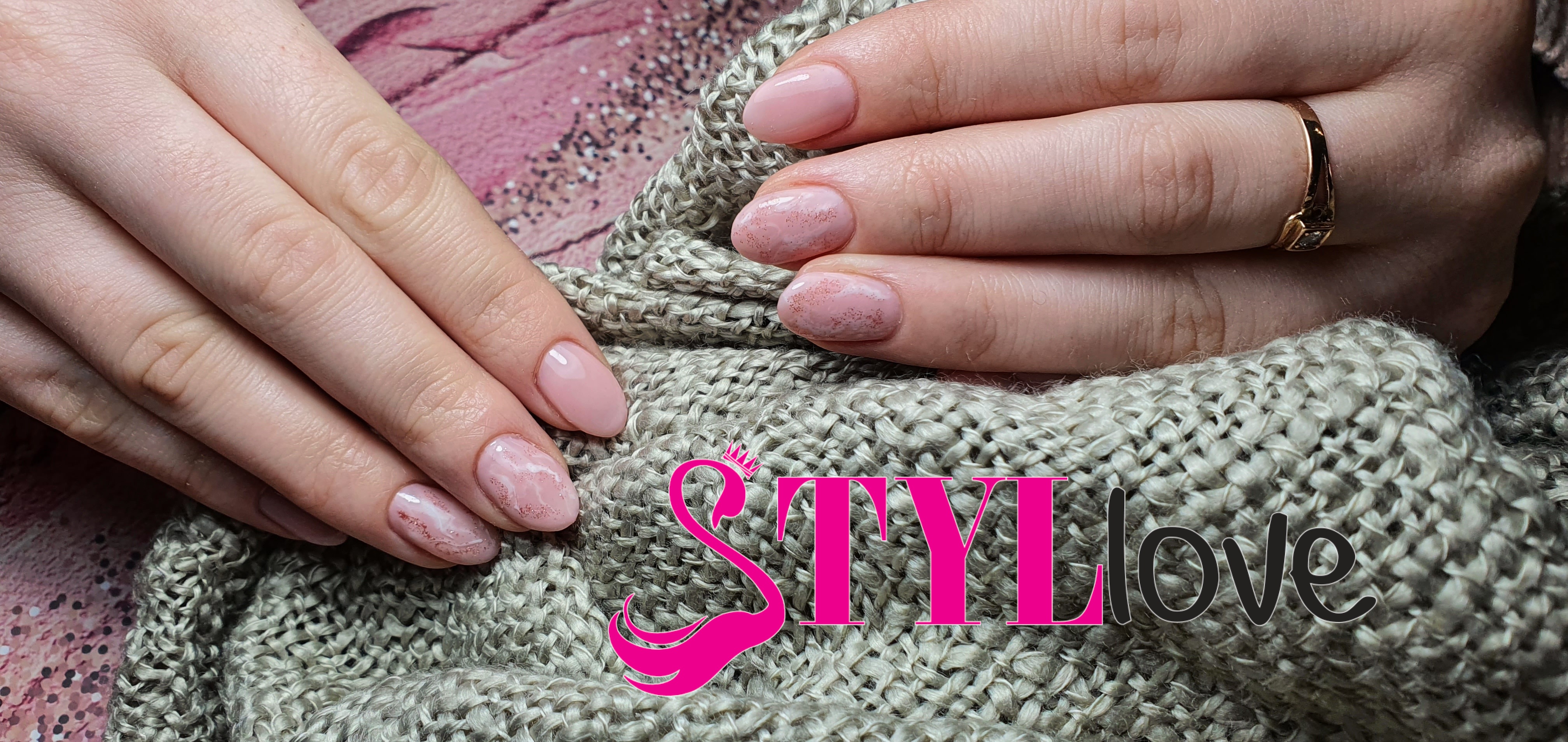 Delikatny, różowy manicure hybrydowy z efektem marmuru i drobnym brokatem na tle grubo plecionego swetra; na palcu złoty pierścionek.