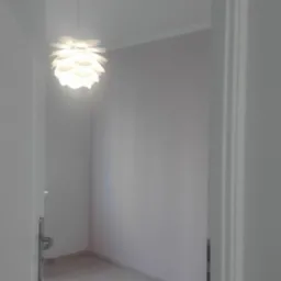 Pomalowane na jasny odcień pokój z plastikową folią ochronną na podłodze widziany zza otwartych drzwi, z wiszącą lampą w kształcie kwiatu.